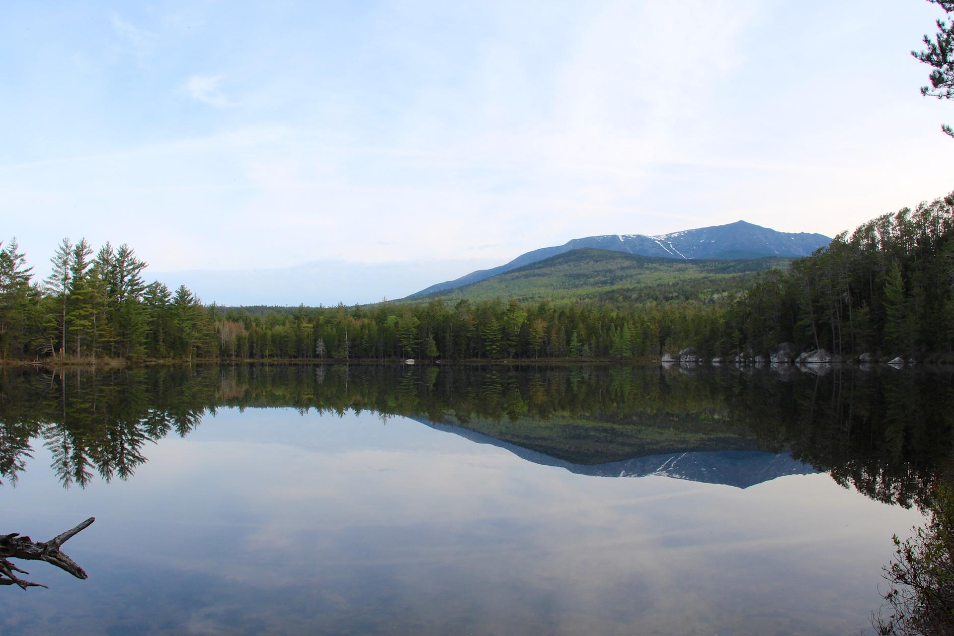 Calm lake reflecting a forested mountain with snow patches under a bright sky.