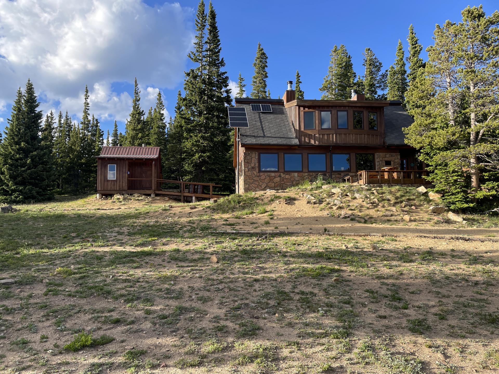 A large cabin and small shed in a clearing surrounded by pine trees.