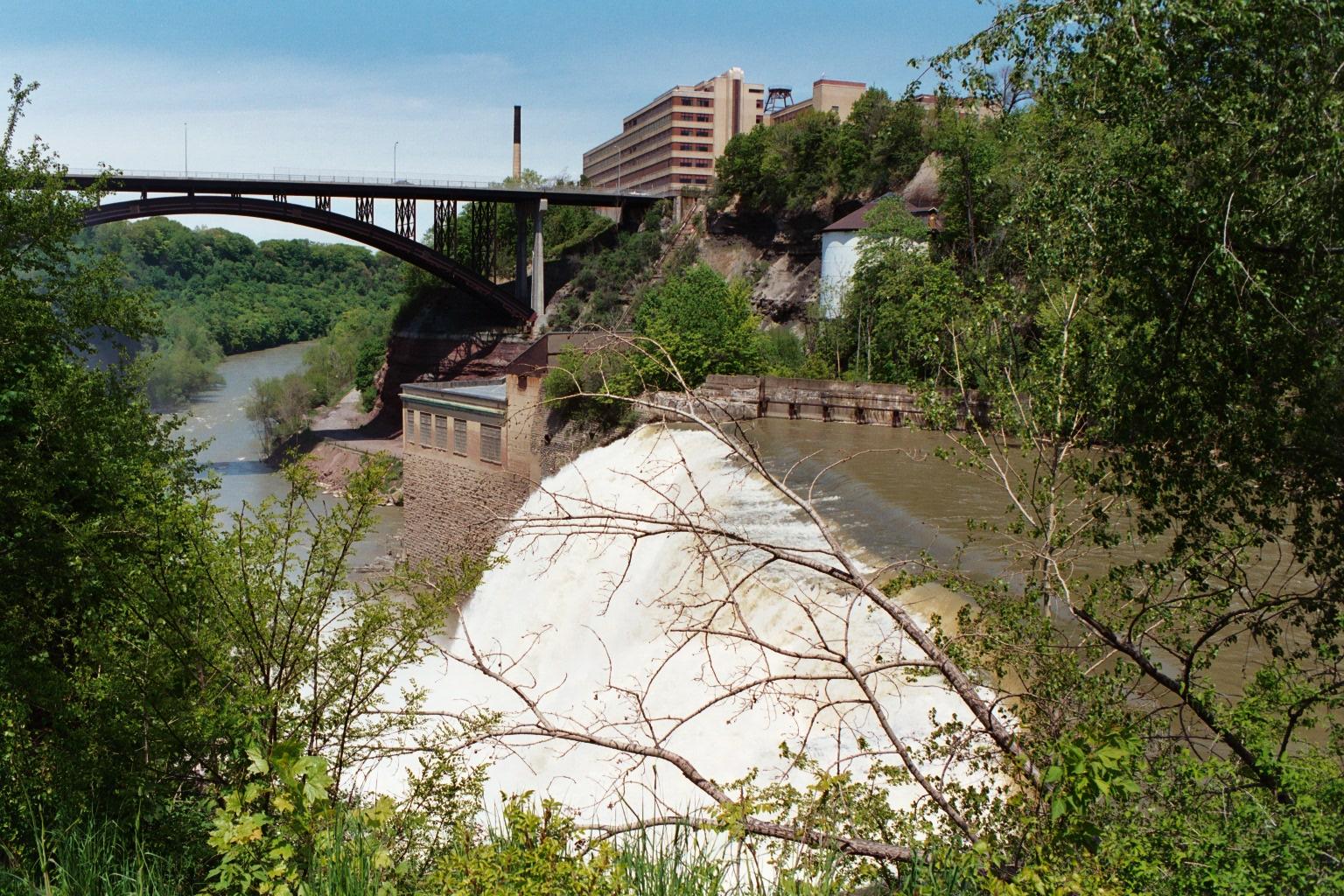 A wide waterfall flows into a river below a bridge and distant buildings.