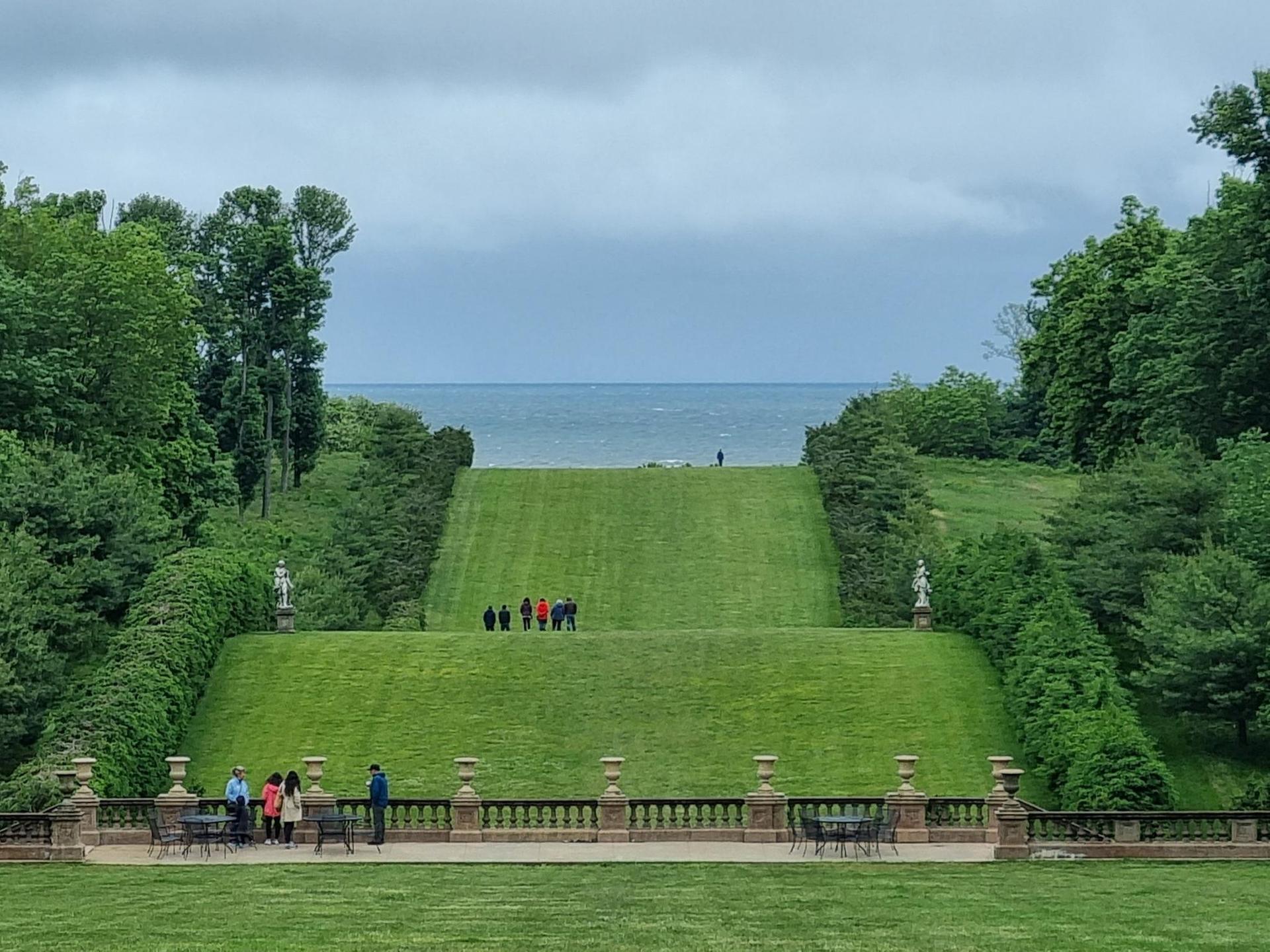 People walk on a vast green lawn sloping towards distant water under an overcast sky.