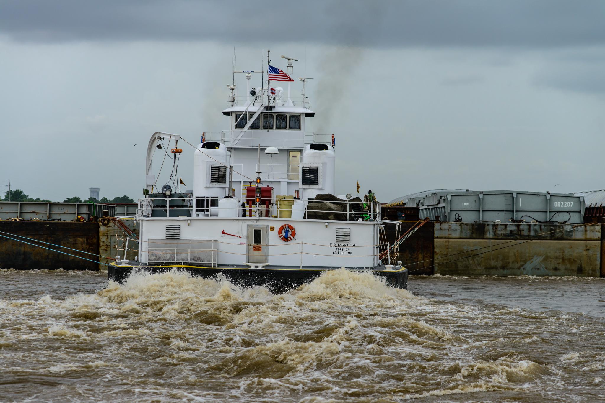 White tugboat pushing barges, creating a large wake in murky water.