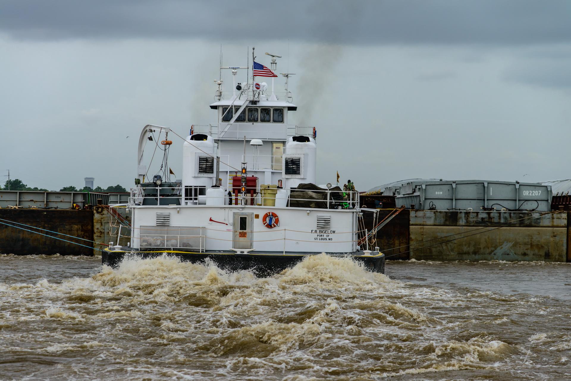 White tugboat pushing barges, creating a large wake in murky water.