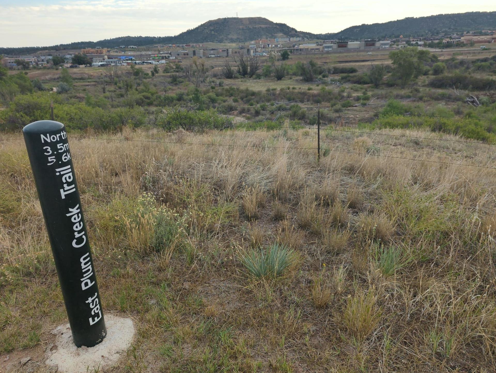 Black trail marker post in a dry, grassy field with distant hills.