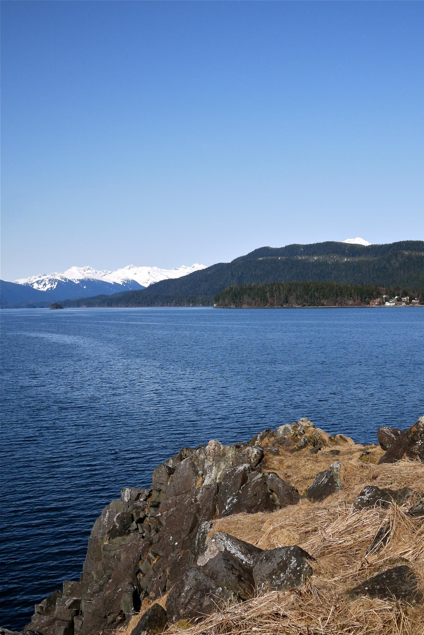 Vast blue water, distant snow-capped mountains, and a rocky foreground under clear skies.