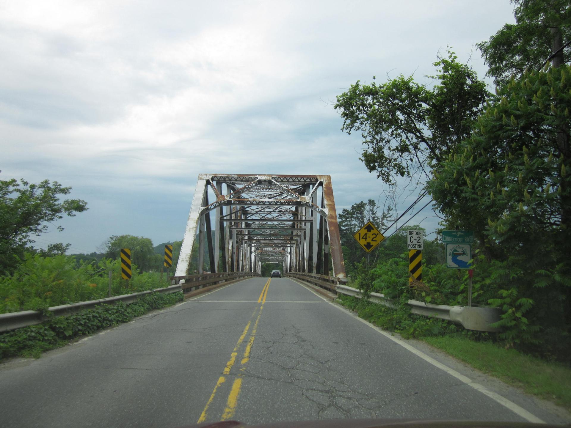 Driving towards a long, metal truss bridge surrounded by green trees.