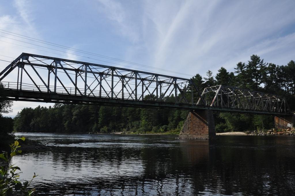 A large metal truss bridge spans a wide river under a partly cloudy sky.