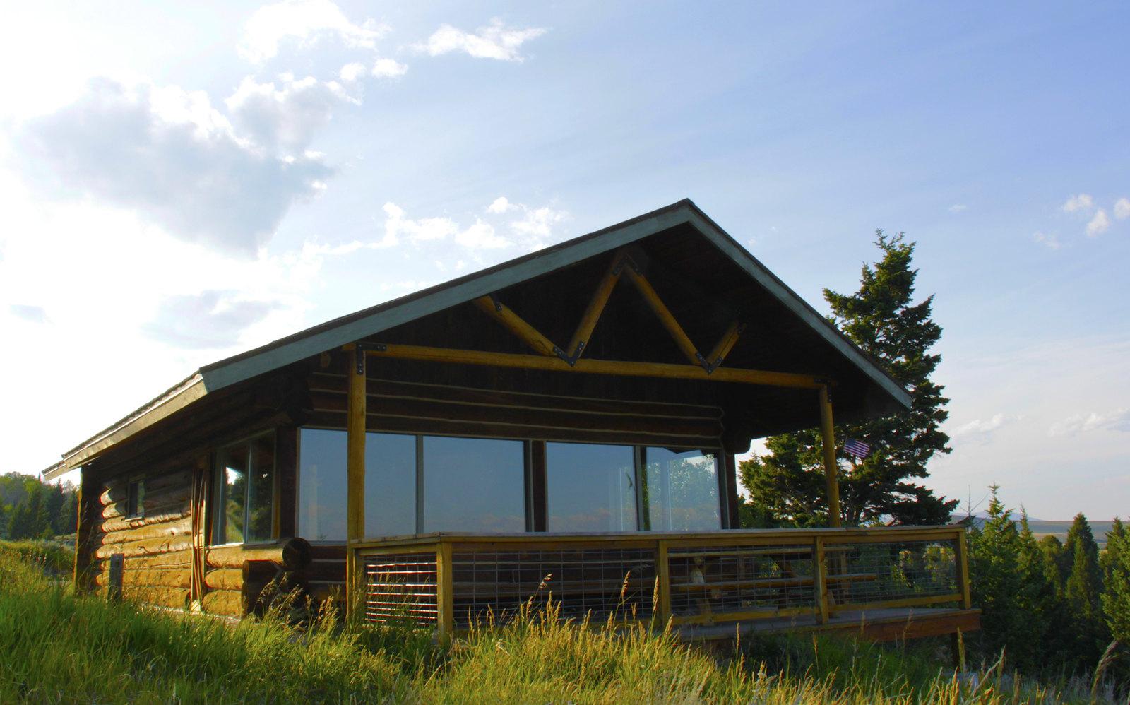 Sunny wooden cabin with large windows and a deck on a grassy hill.