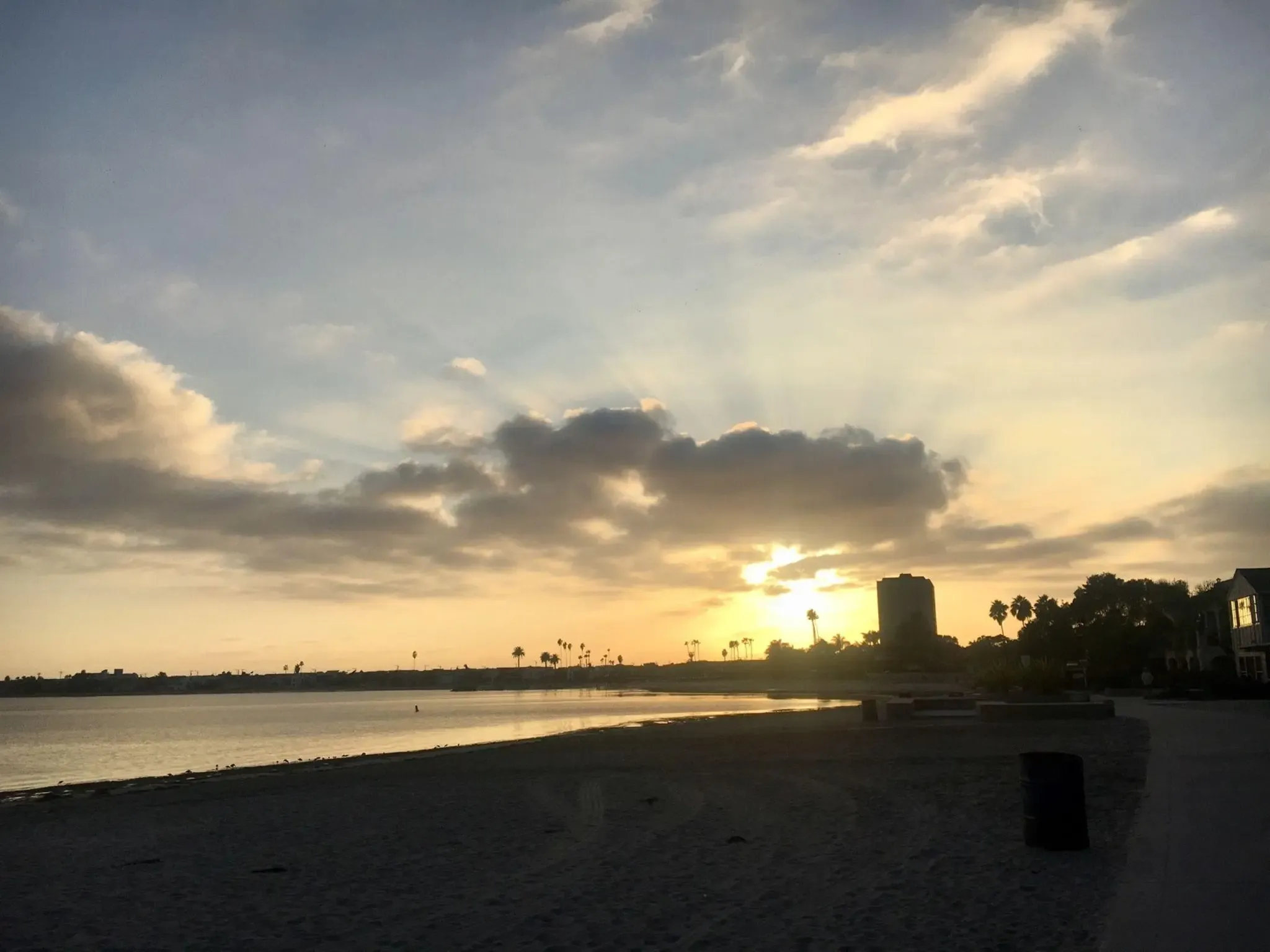 Golden sunset over a calm bay with a sandy beach and distant buildings. - Mission Bay Bike Path