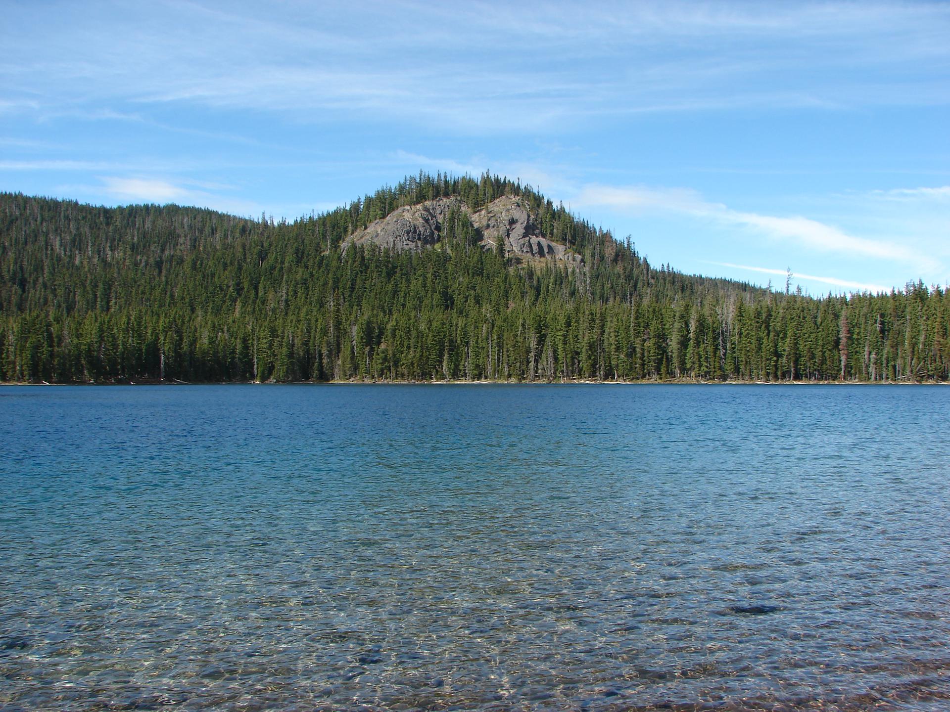 Clear lake water in foreground, with a rocky, forested hill under blue sky.