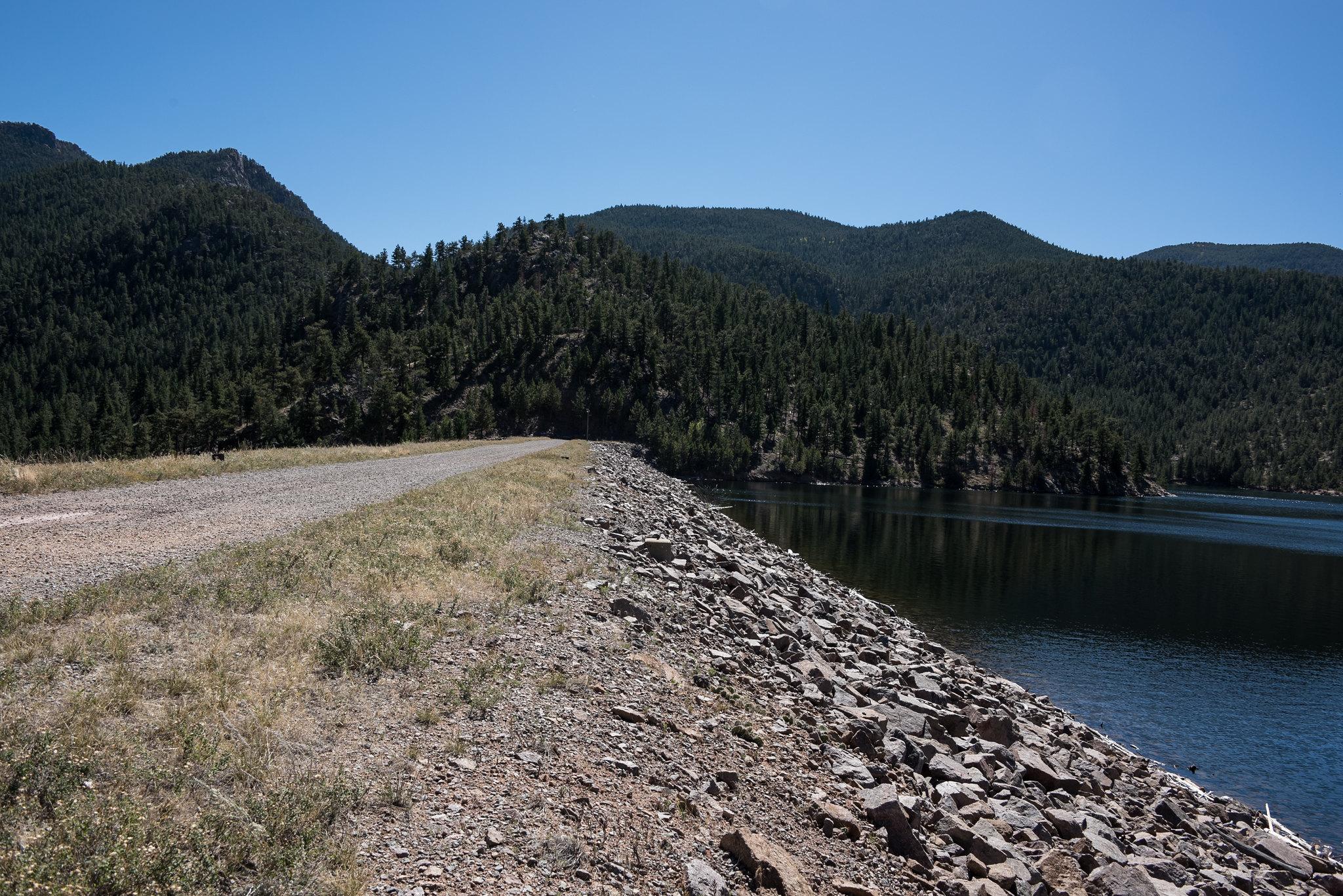 A rocky dam wall next to a calm lake and forested mountains.