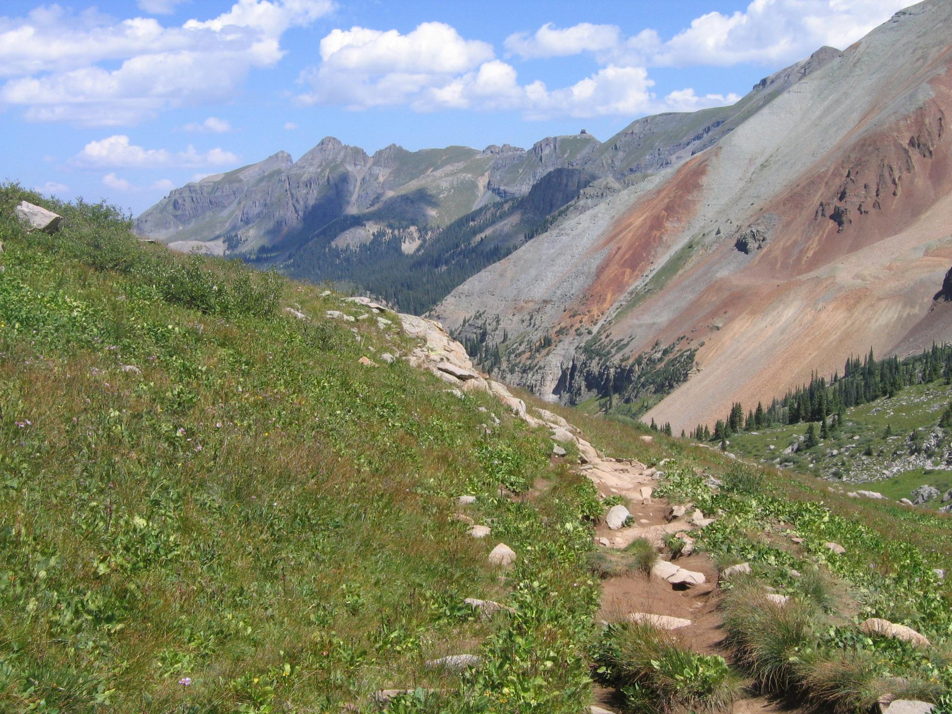 Sunny mountain landscape with a rocky trail through green grass and colorful peaks.