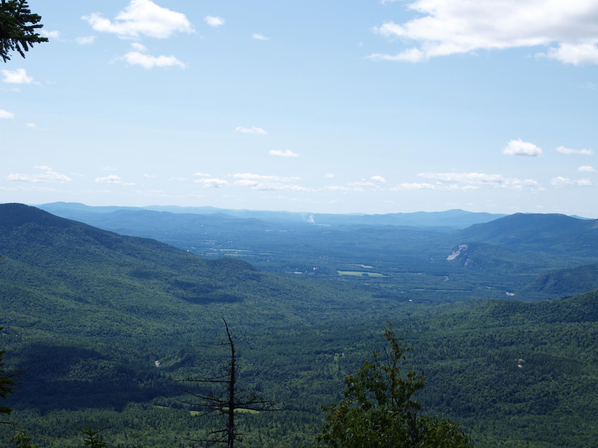 Vast forested mountain landscape under a bright blue sky with scattered clouds.