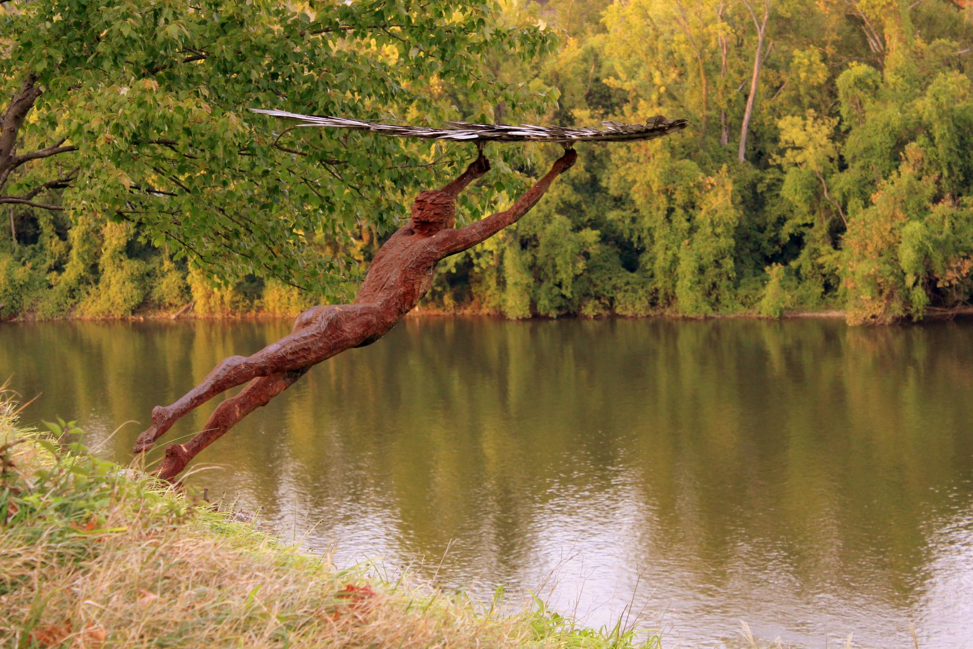 Sculpture of a person reaching over a calm river with lush green banks.