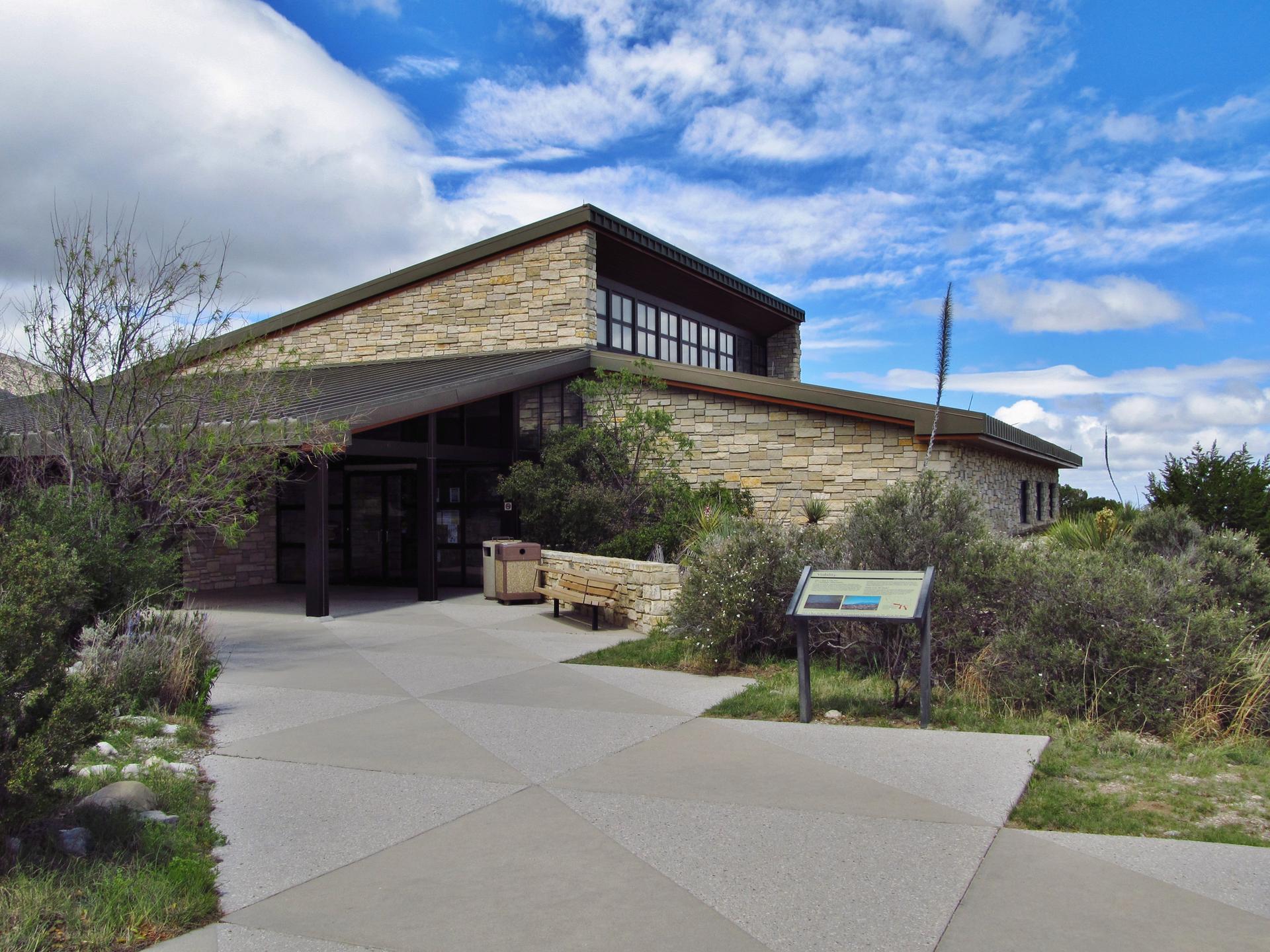 Modern stone building with large windows, surrounded by green bushes under blue sky.