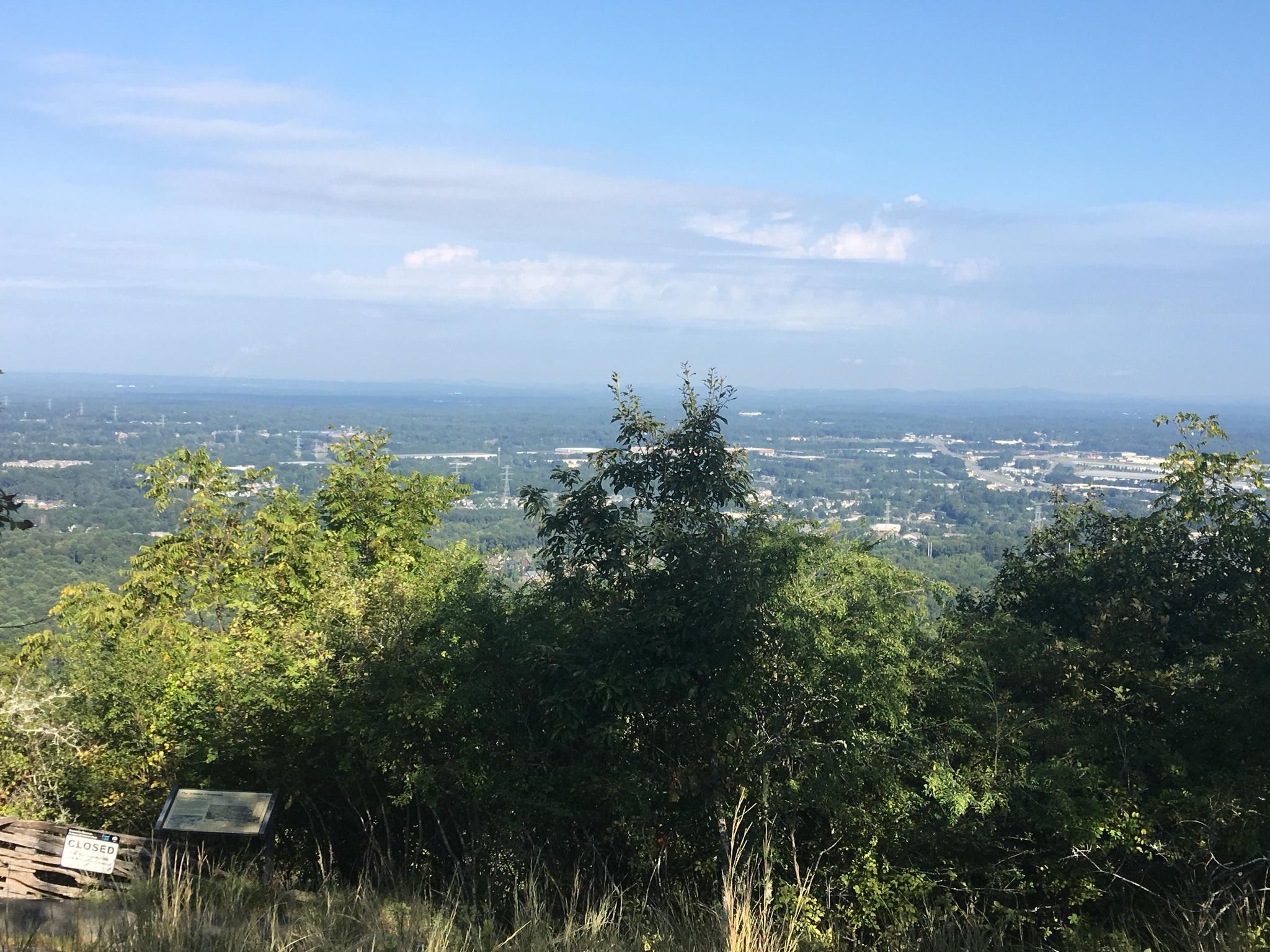 Mountain overlook with distant city, green trees, and blue sky.