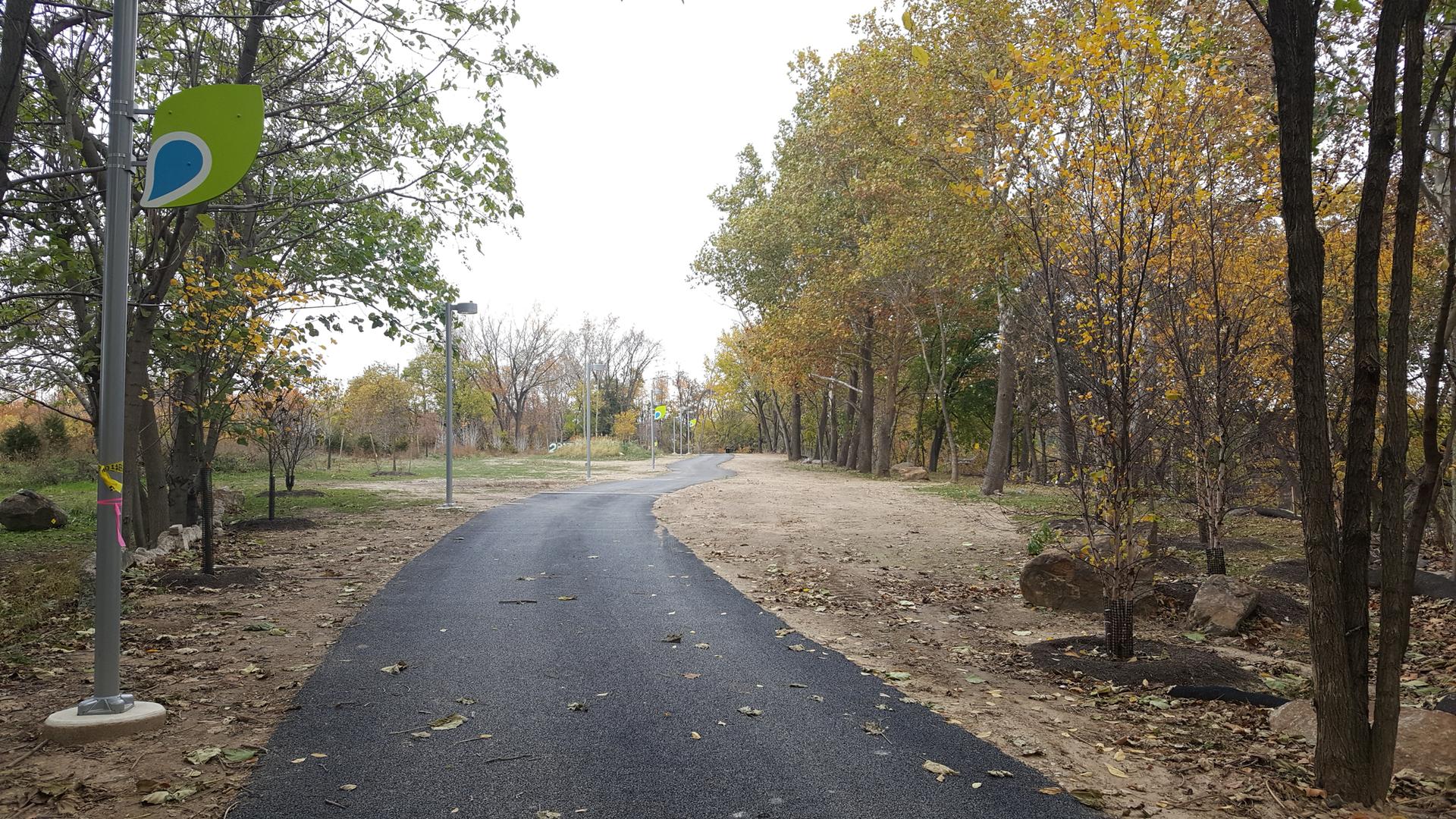 Winding paved path through trees with autumn foliage under an overcast sky.