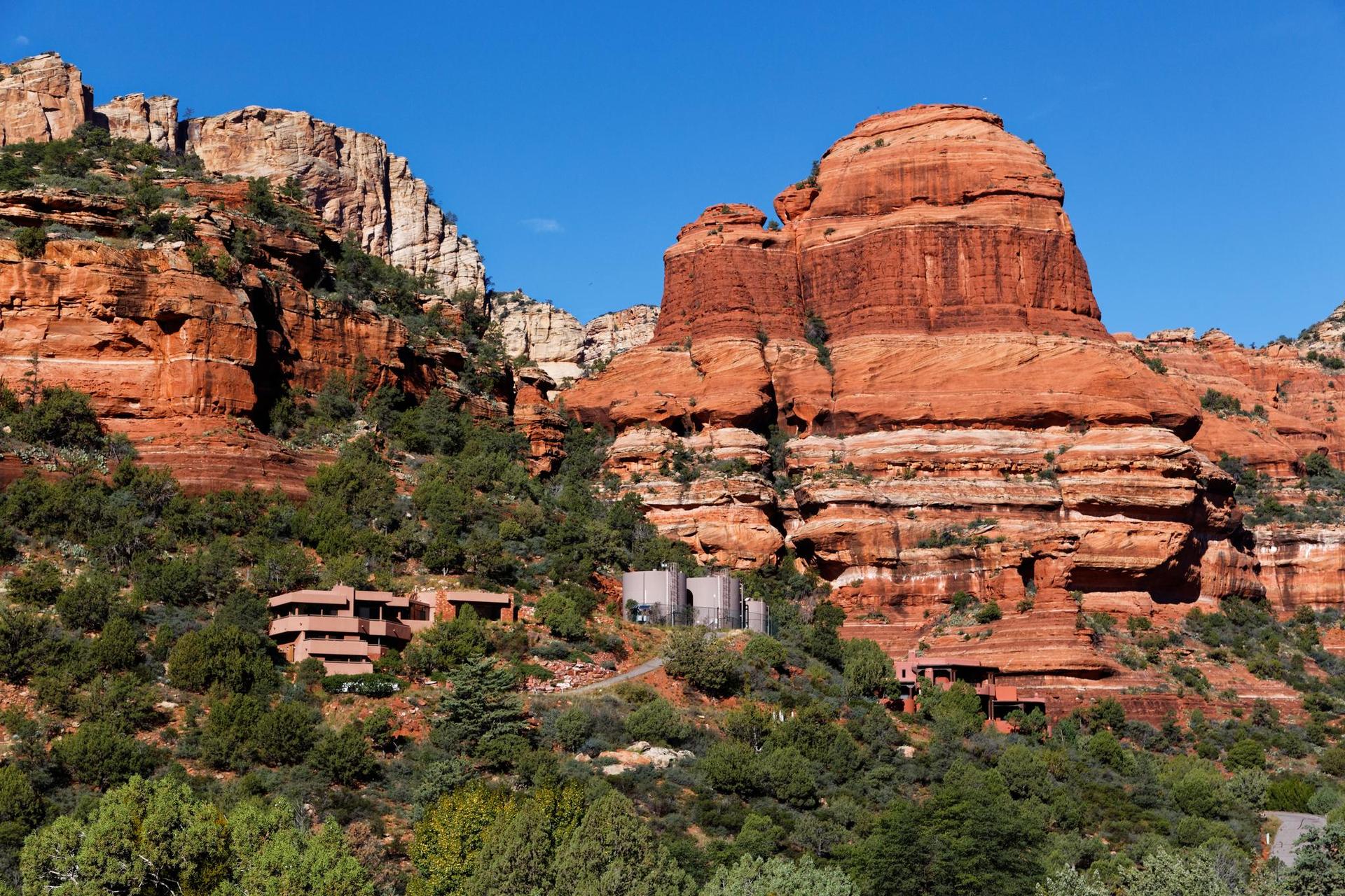 Majestic red rock formations rise above buildings surrounded by green trees.