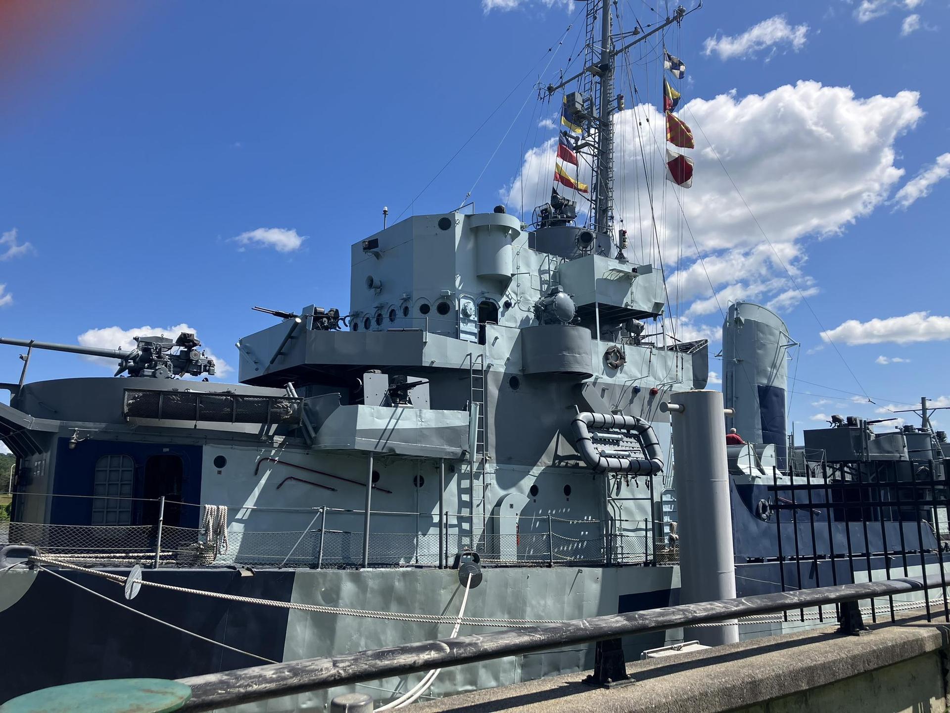 Large gray warship docked, flags flying, under a blue sky.
