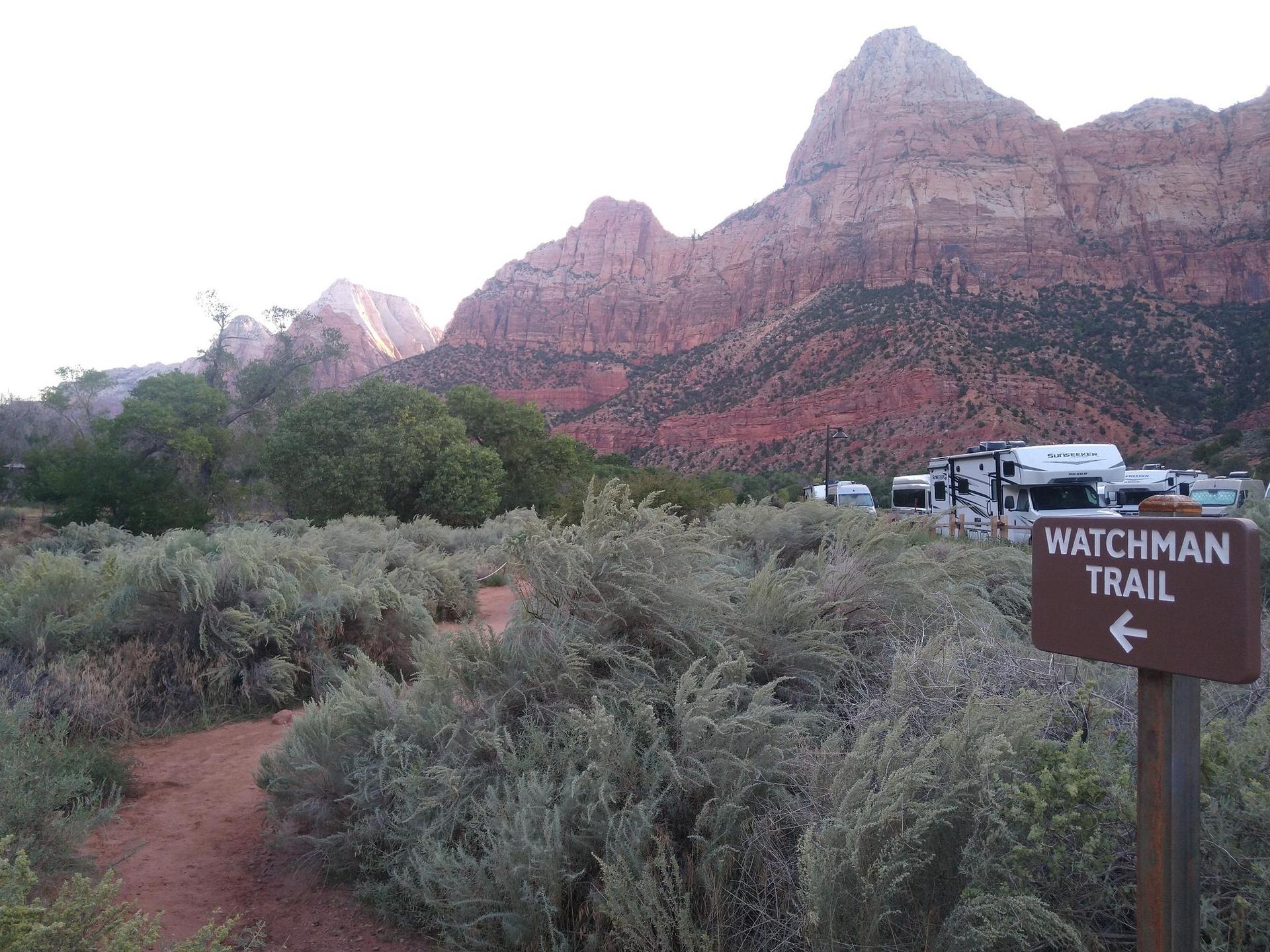 A trail sign points left through desert shrubs with RVs and mountains in the background.