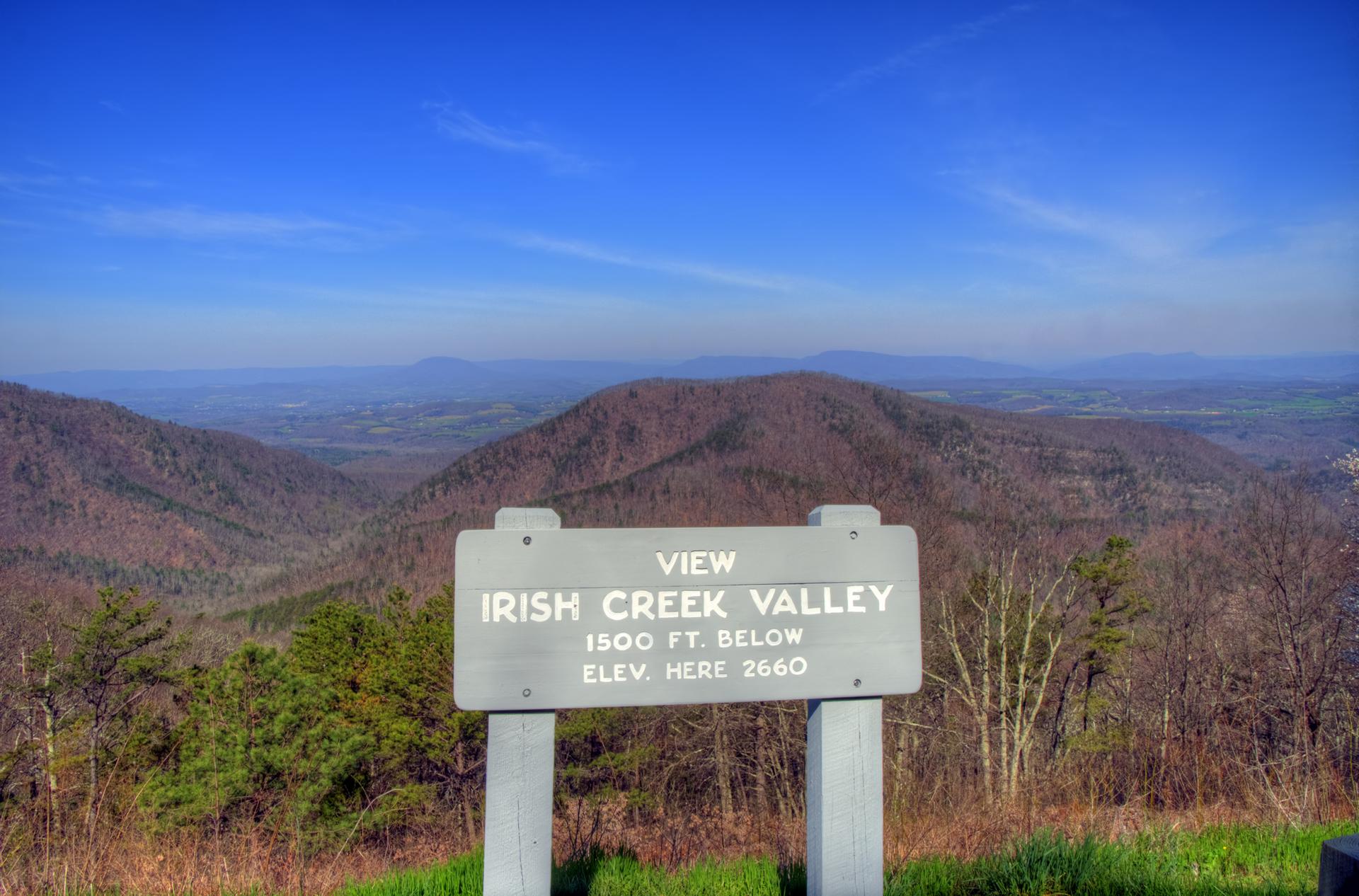 A wooden sign overlooks a vast, hazy mountain valley under a clear blue sky.