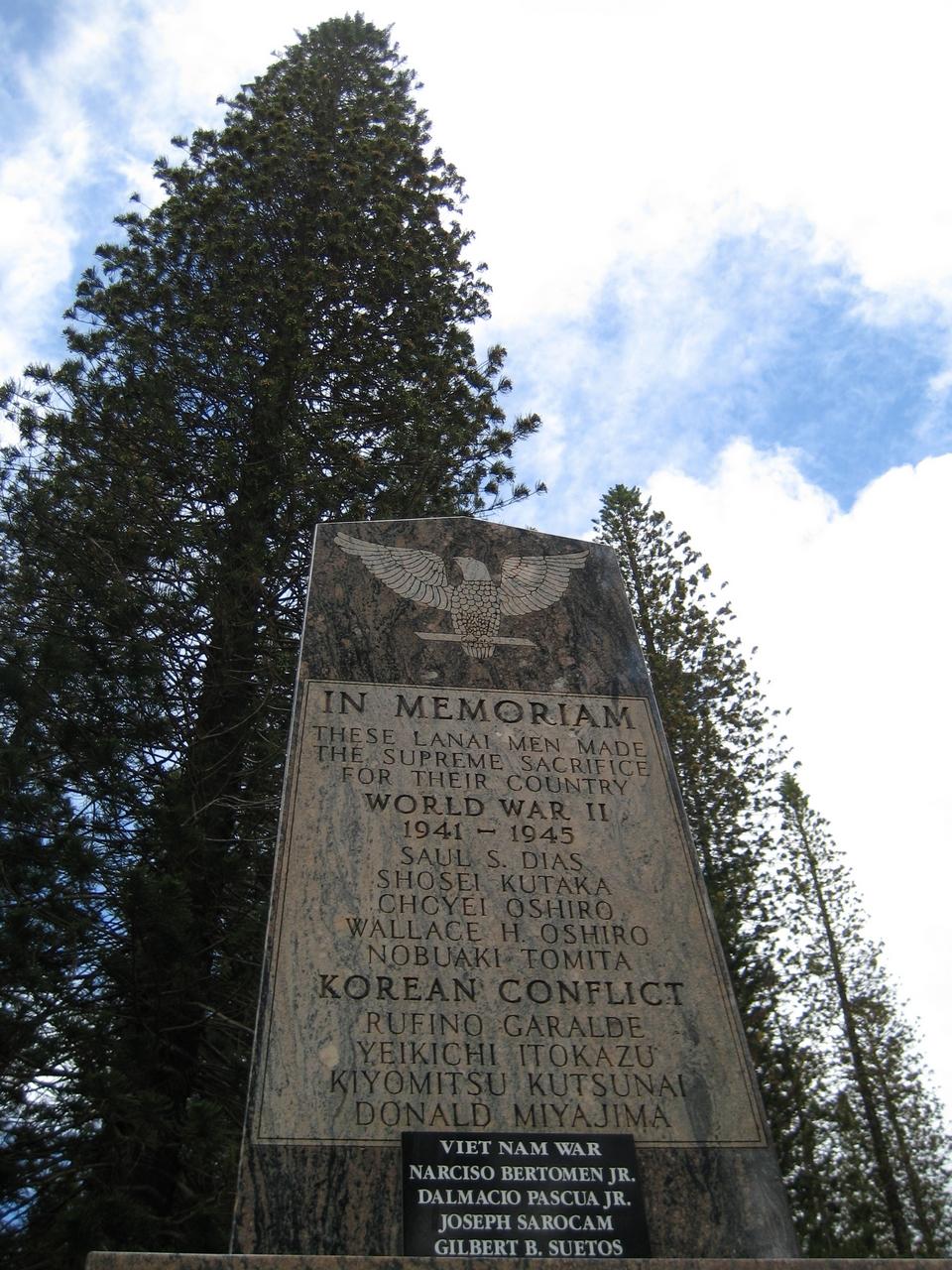 Stone memorial monument under tall pine trees against a partly cloudy sky.