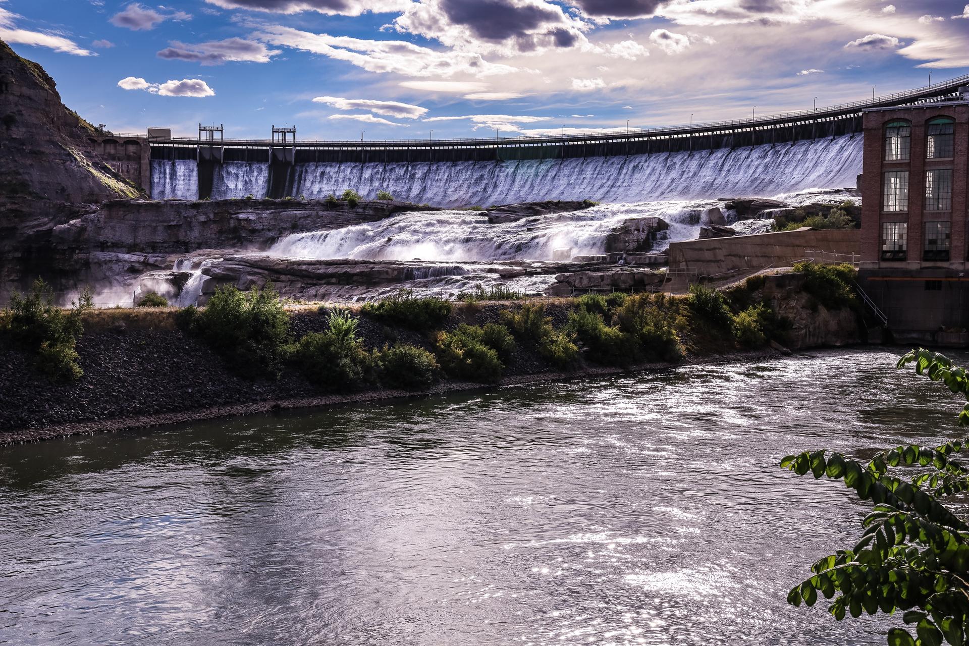 Large dam with water cascading over, flowing into a wide river under a cloudy sky.