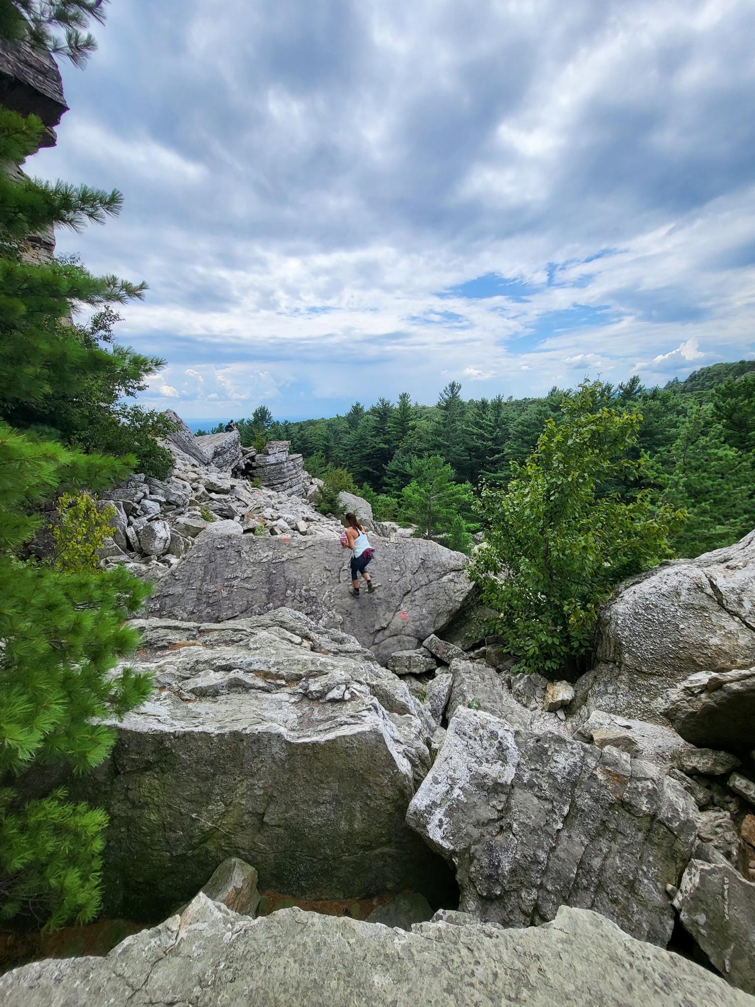 Person scrambling on large boulders under a cloudy sky, surrounded by forest.