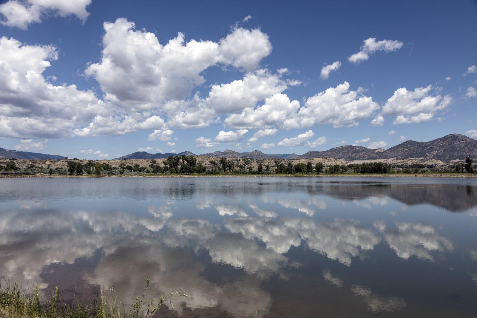 A serene lake reflects a bright blue sky with white clouds and distant mountains.