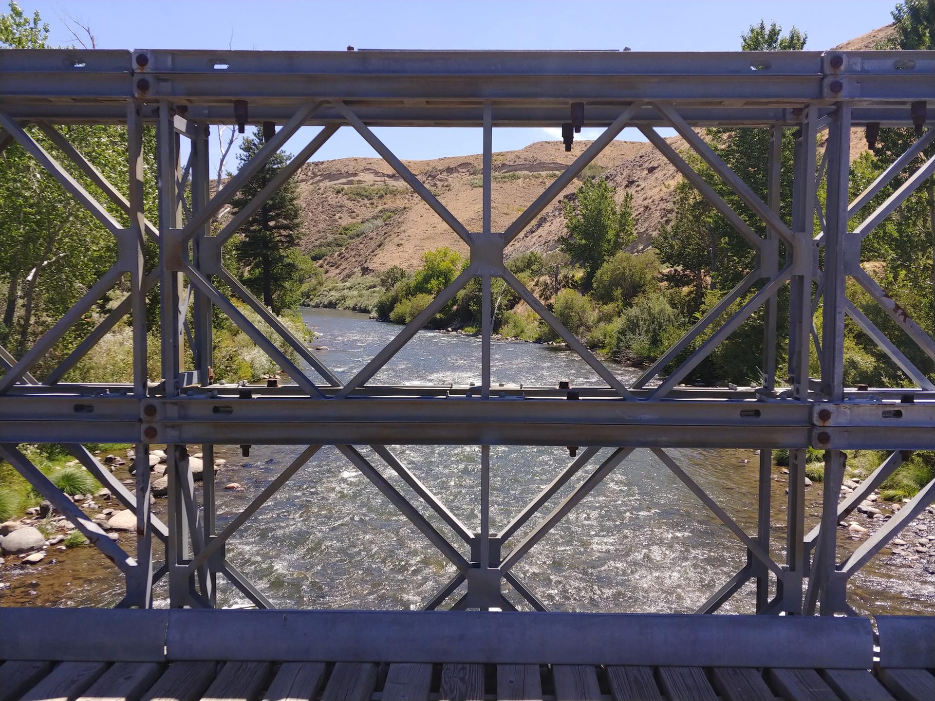 View through a metal bridge structure of a river and distant hills.