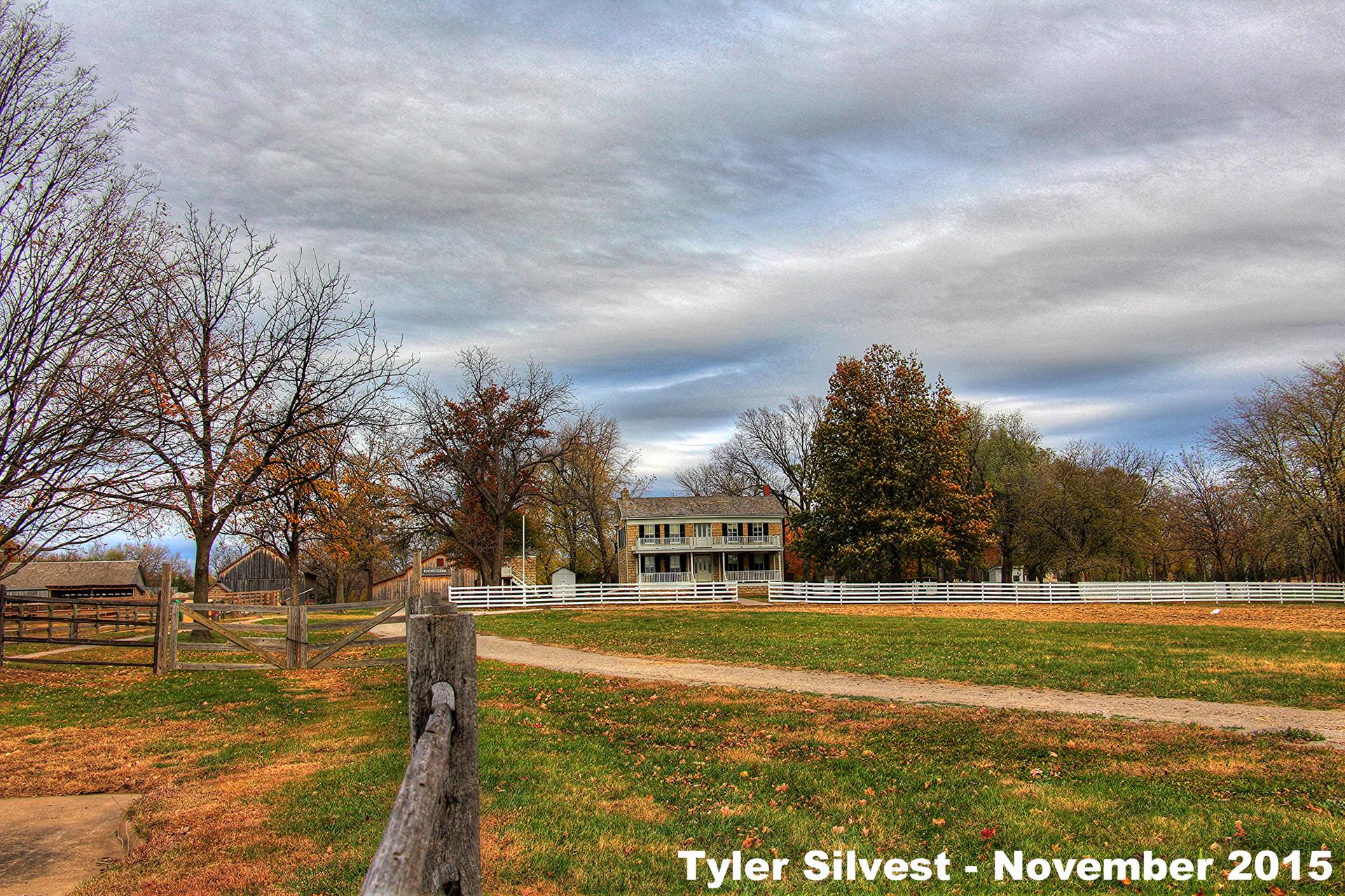 Historic house behind a white fence, surrounded by autumn trees under cloudy sky.