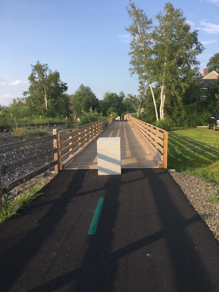 Paved path leading to a wooden bridge under a clear sky, with long shadows.