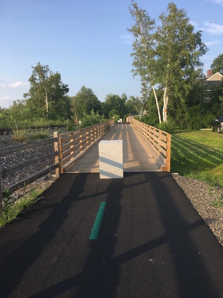 Paved path leading to a wooden bridge under a clear sky, with long shadows. - WOW Trail