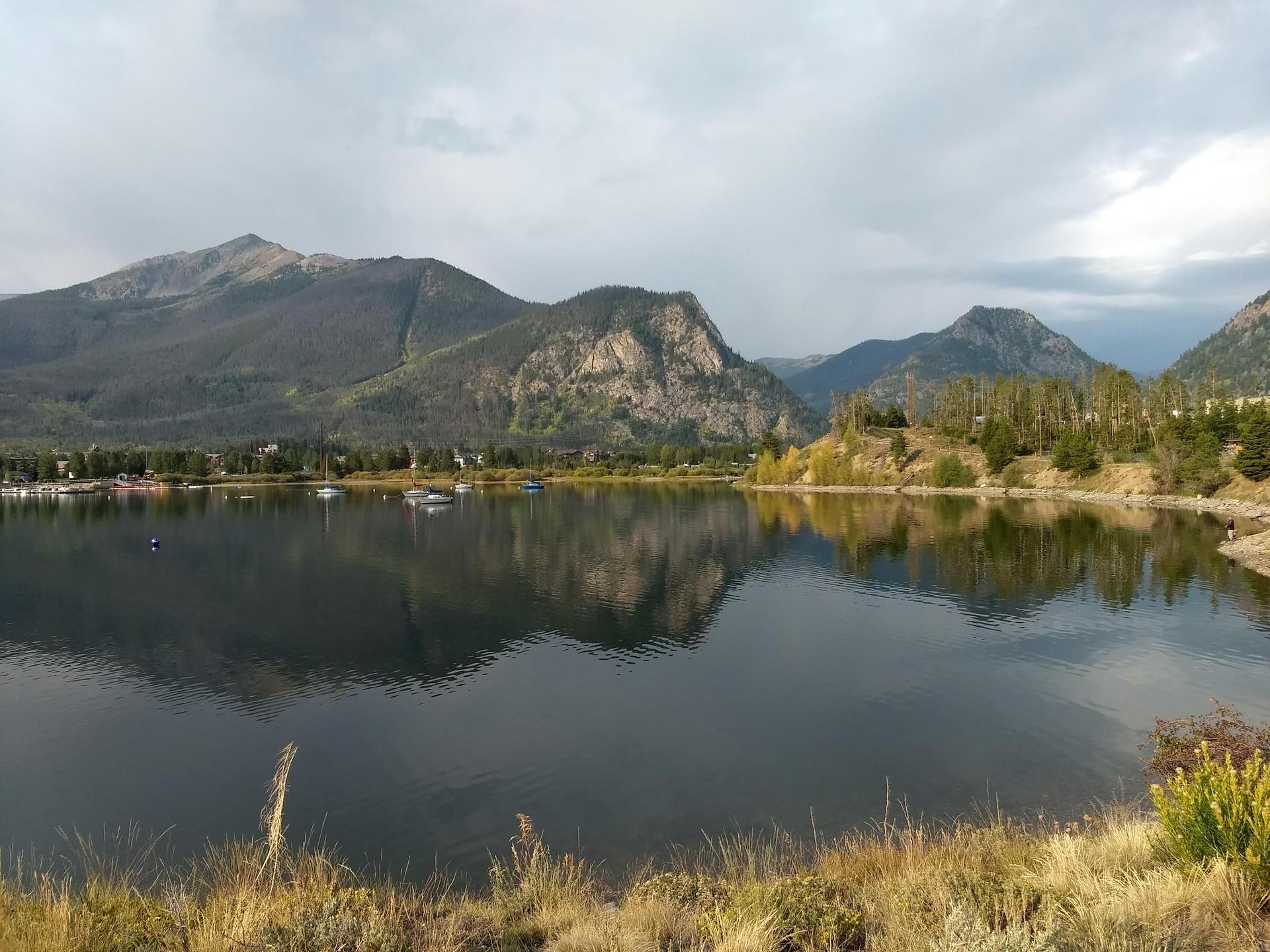Overcast view of a calm lake reflecting mountains, with boats in the distance.