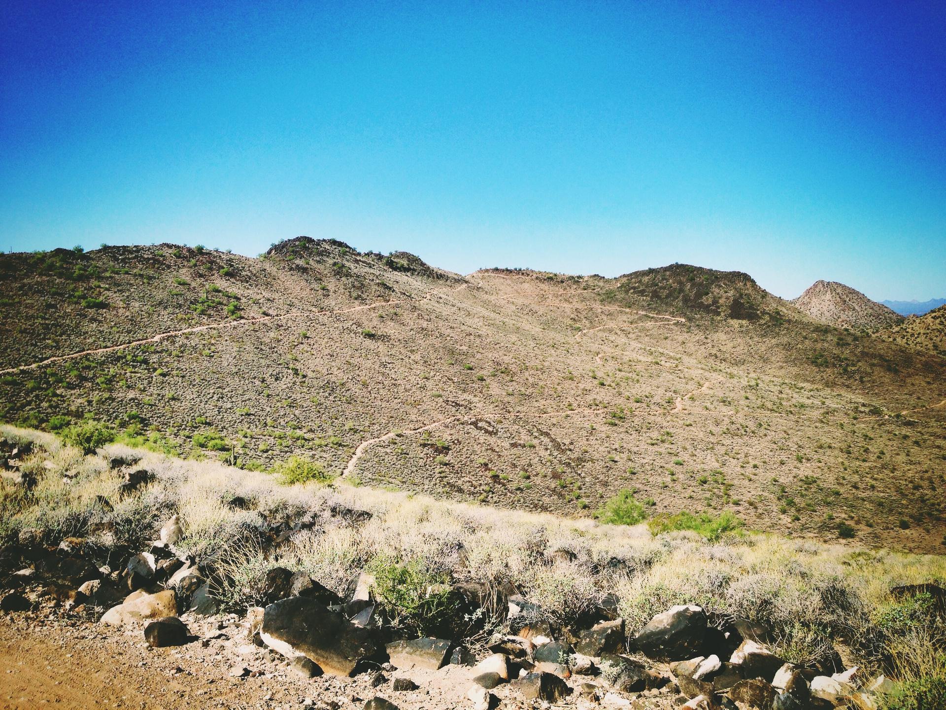 Arid desert hills with a winding trail under a bright blue sky.