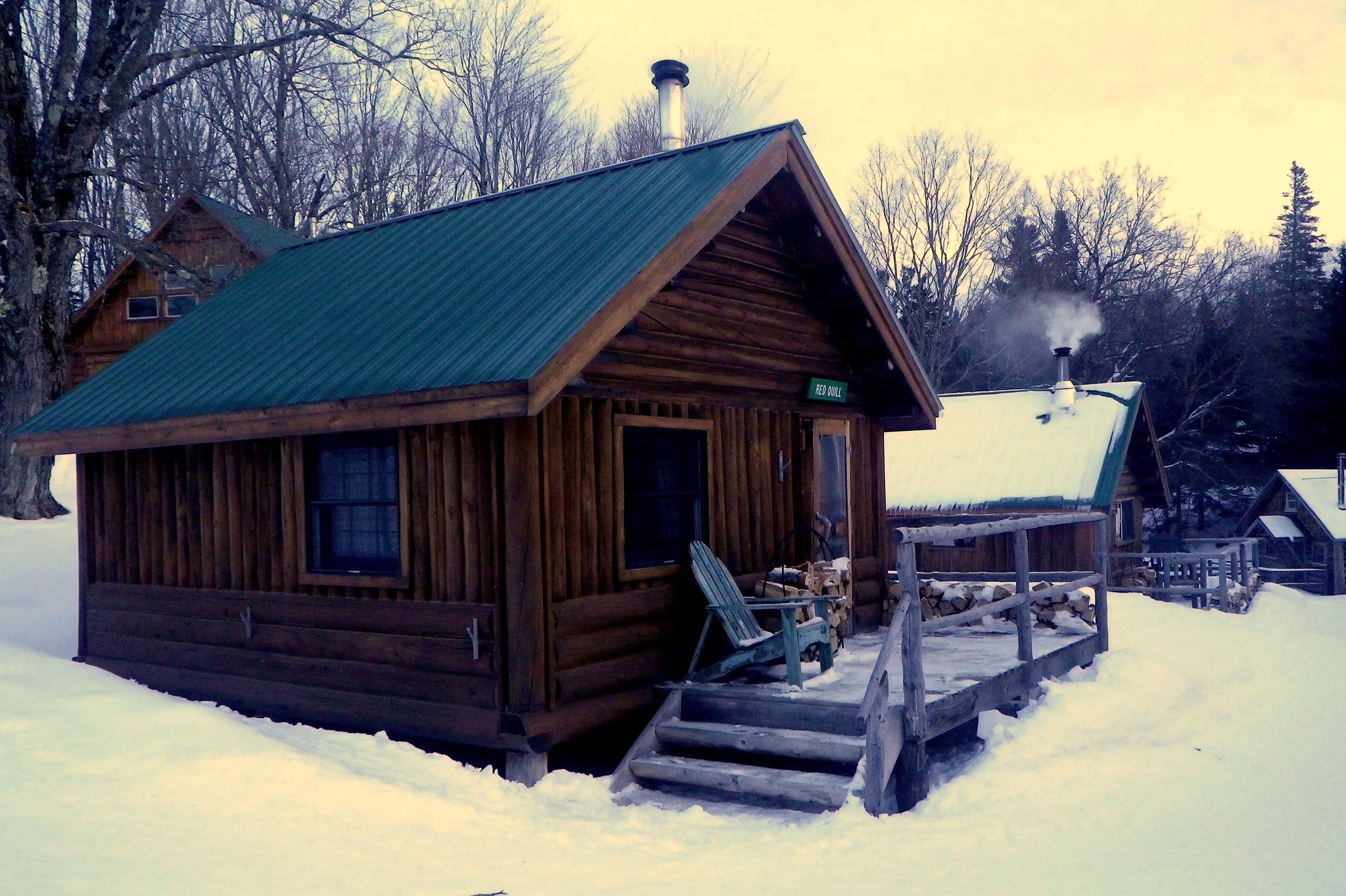 Rustic wooden cabin with green roof, smoke from chimney, in snowy woods. - Lodge to Lodge Trail: Gorman Chairback to Little Lyford