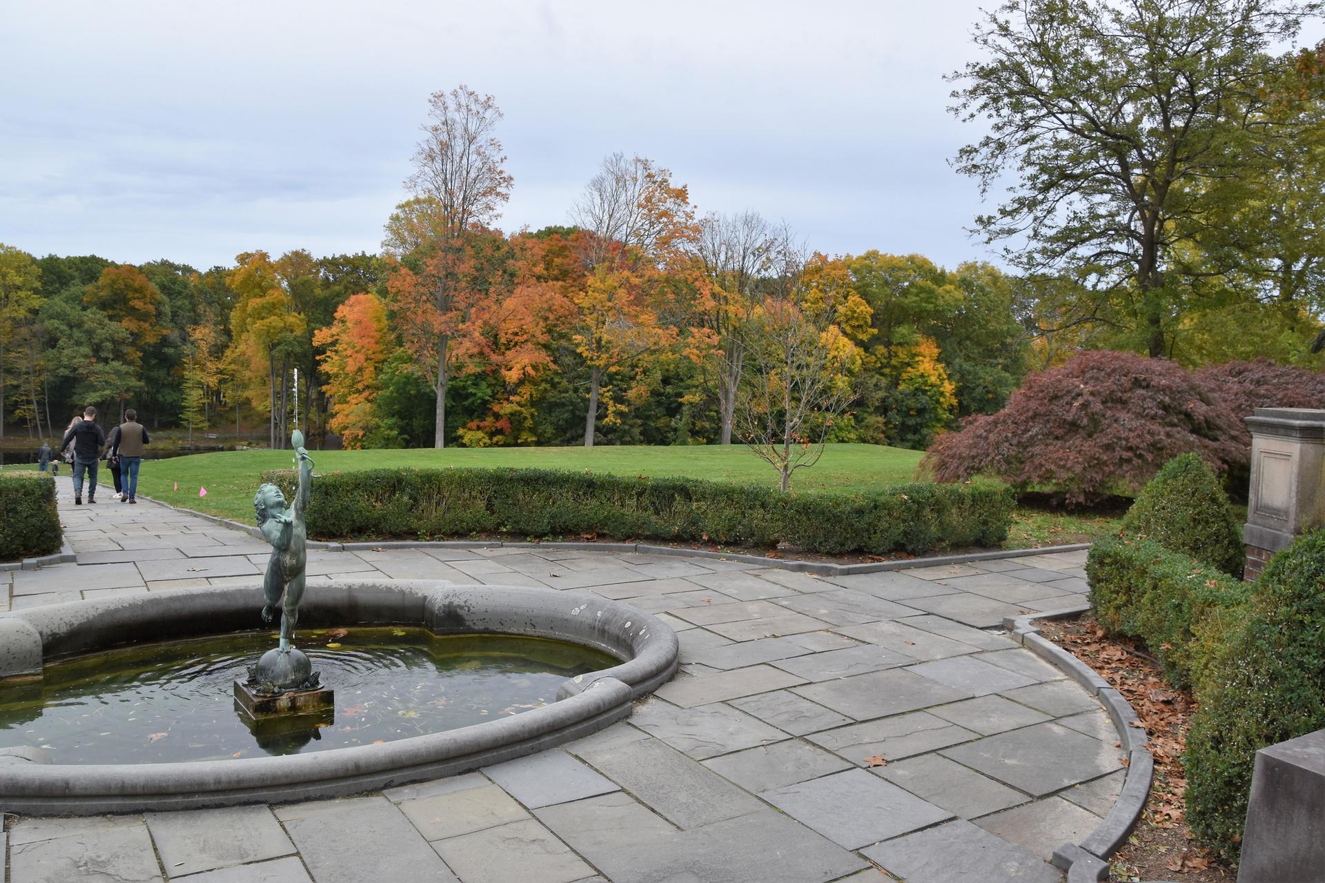 Fountain with child statue, people walking on paved patio, autumn trees under overcast sky.
