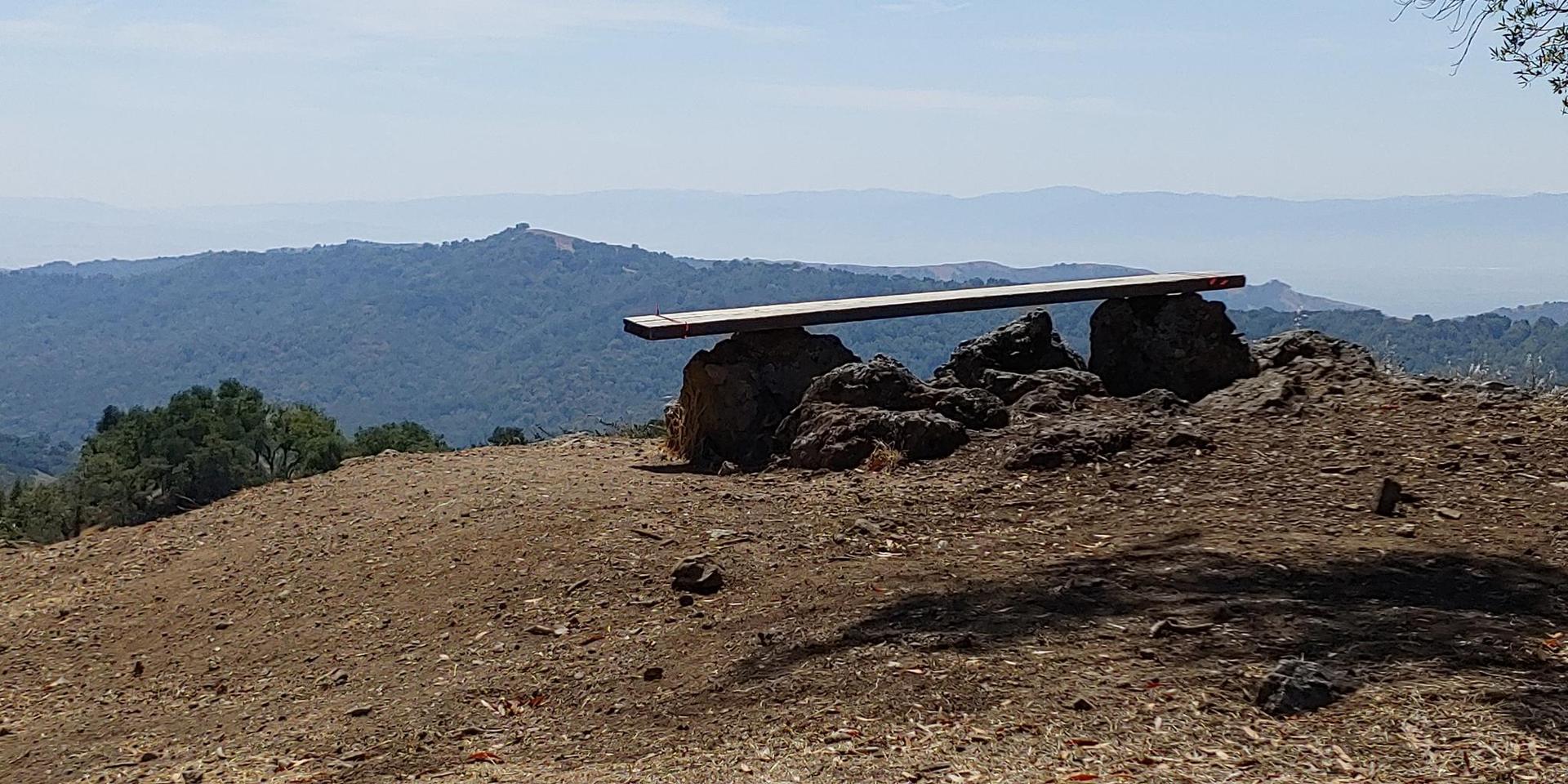 A rustic wooden bench on a rocky hill overlooks hazy distant mountains.