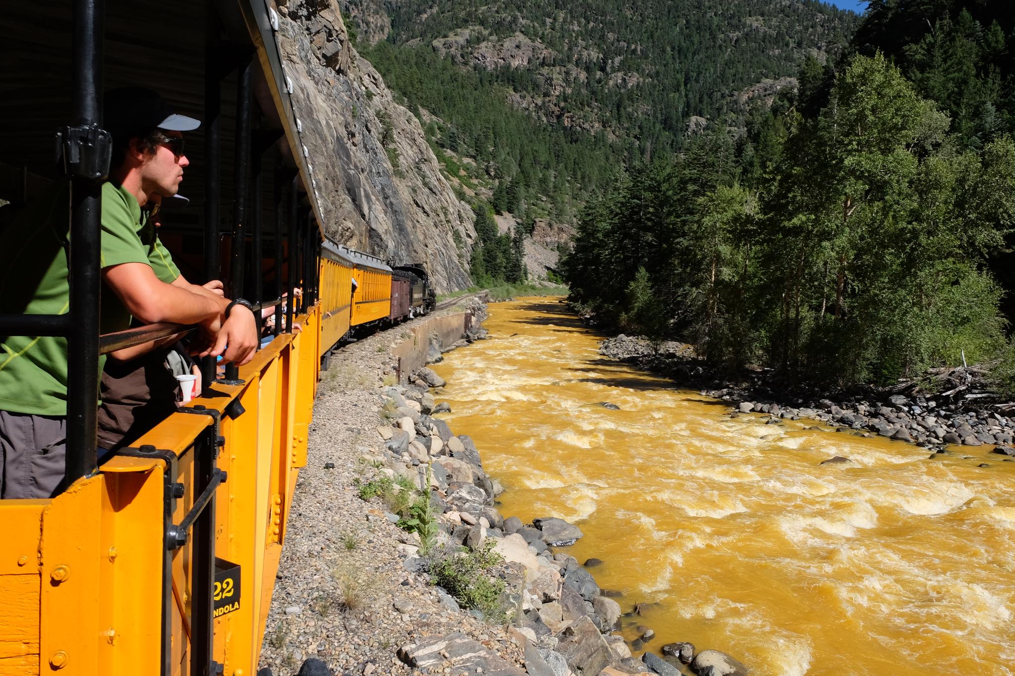 Man looking out from a yellow train car beside a muddy river.