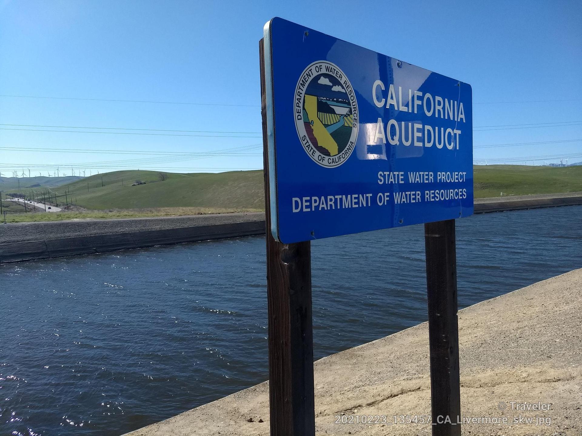 A blue sign stands beside a wide canal with green hills beyond.