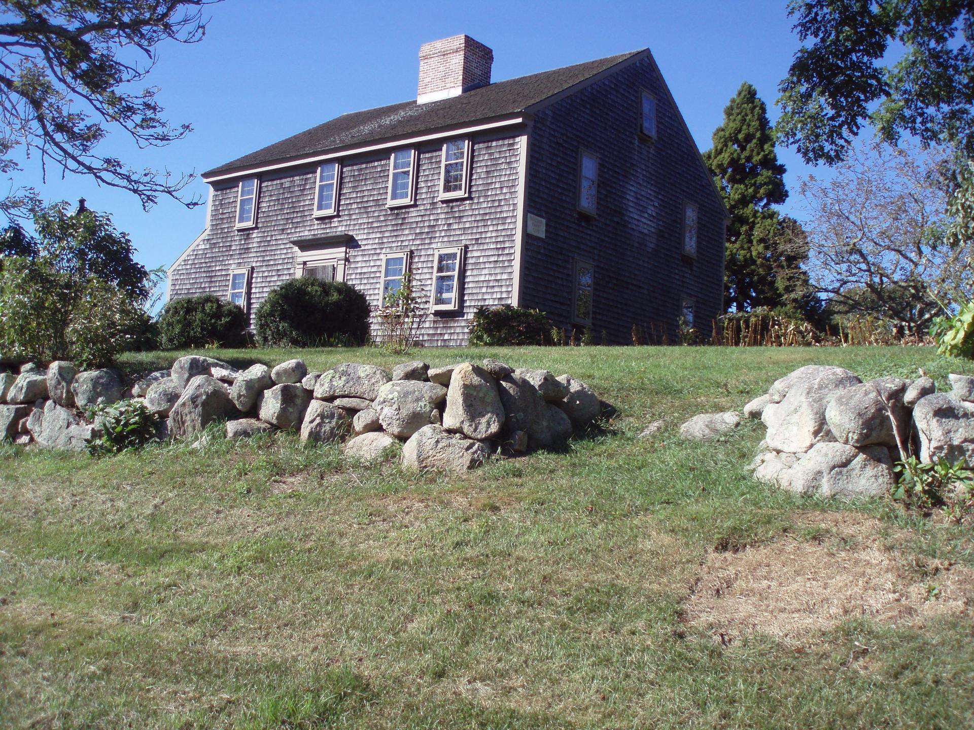 Old shingled house on a sunny grassy hill with a stone wall.