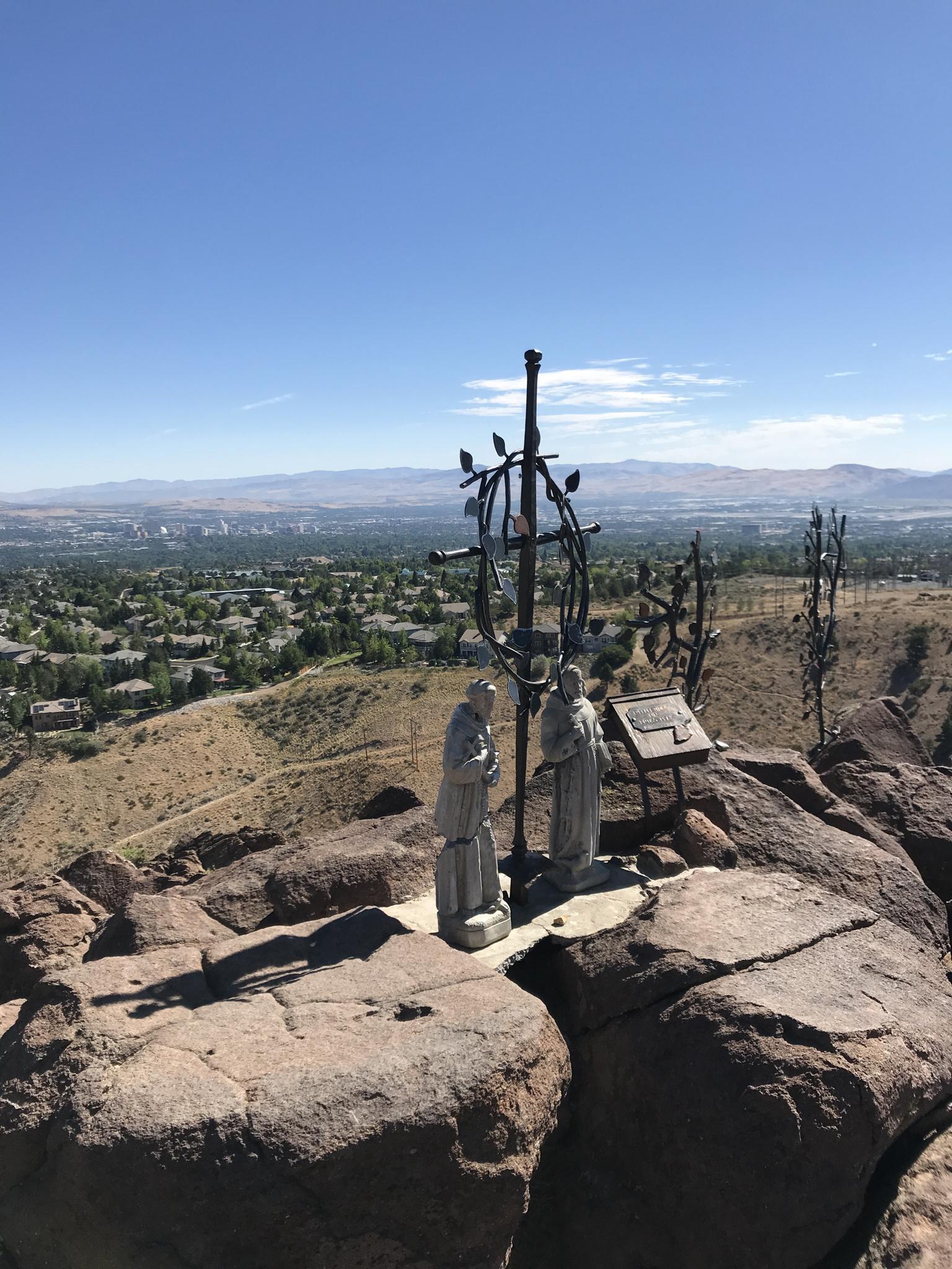 Religious statues and crosses on a rocky peak overlooking a city.