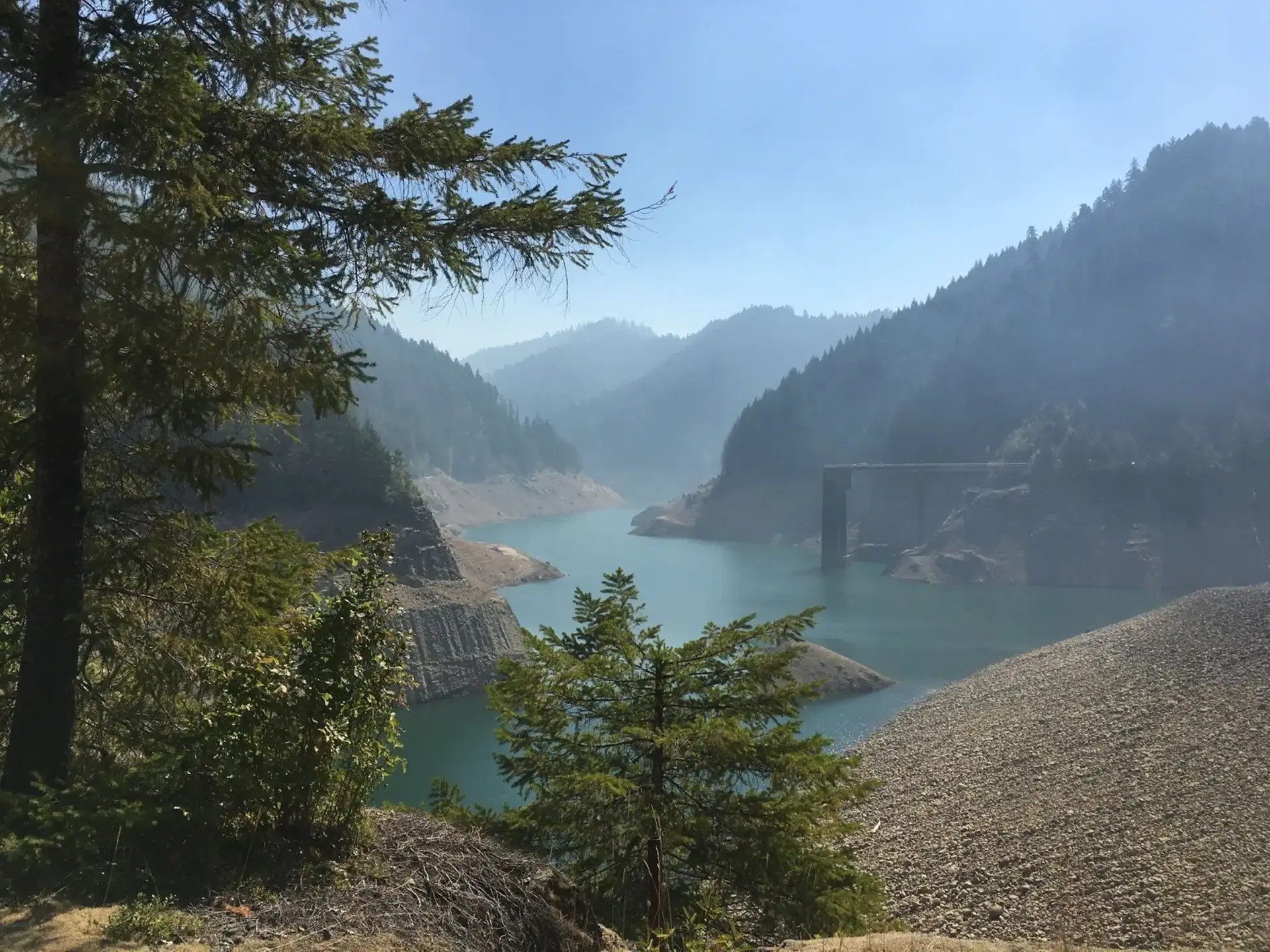 A hazy mountain lake with a bridge and forested slopes under bright skies.