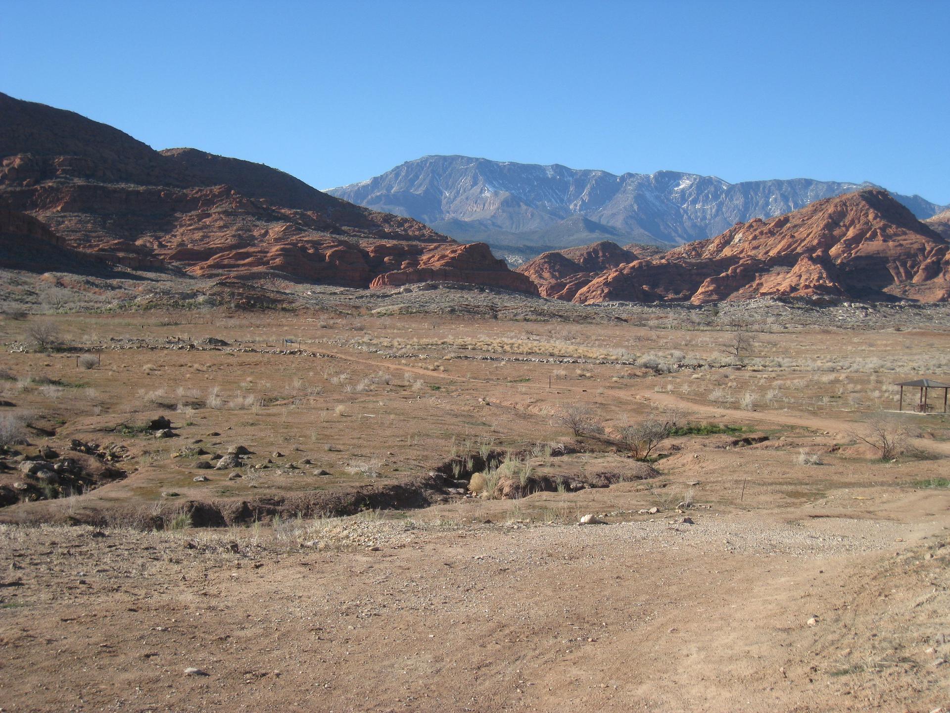 A wide desert valley with red rock formations and distant mountains under blue sky.