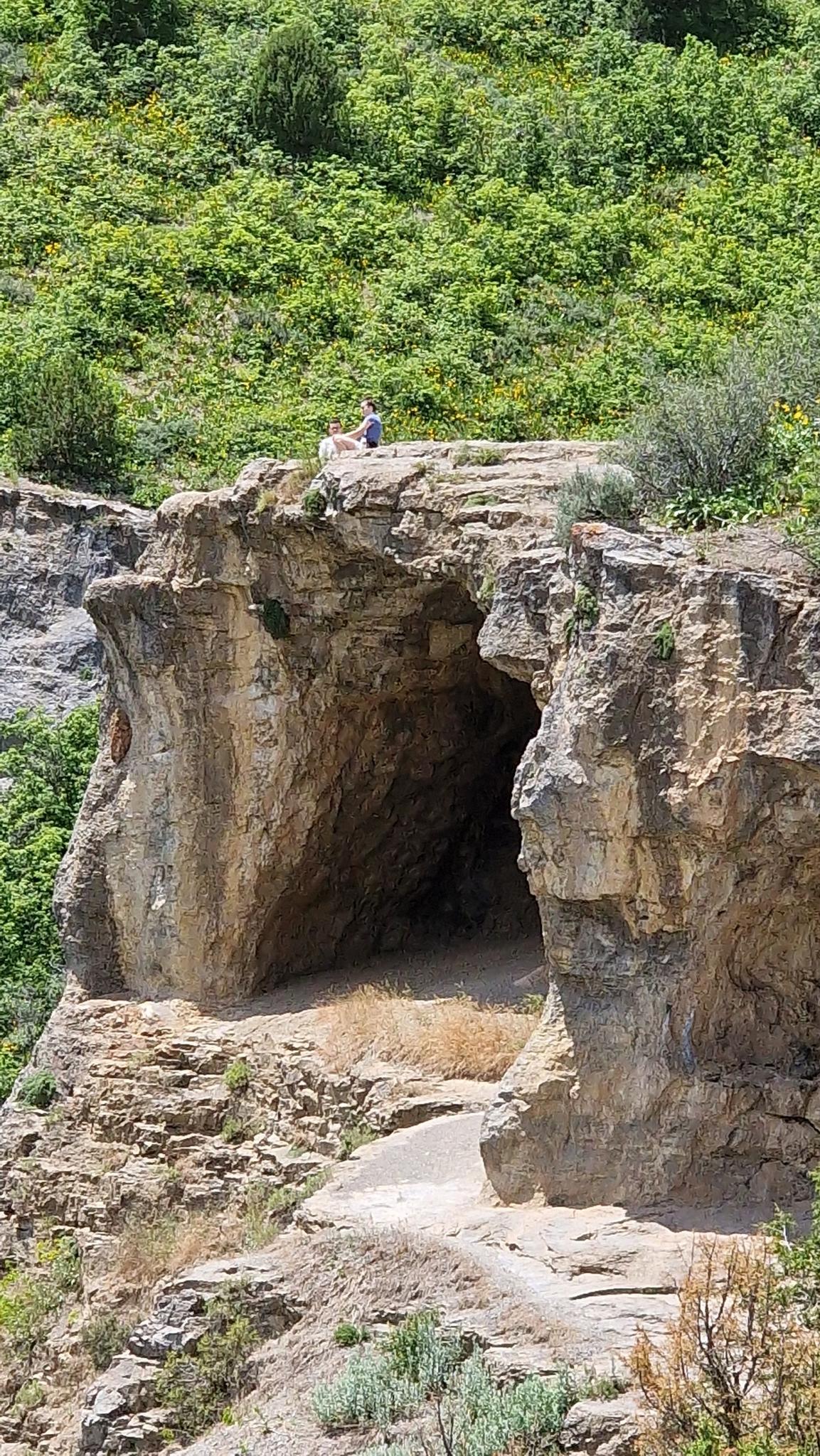 Person sitting atop a large rock formation with a cave entrance below.