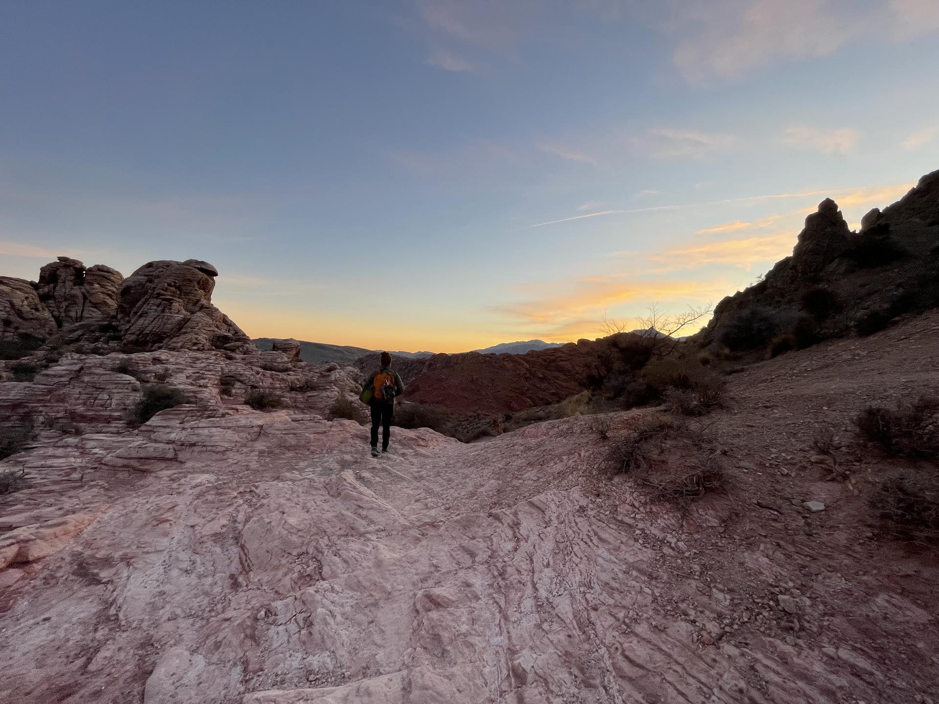 Hiker on rocky terrain facing a sunset over distant mountains.
