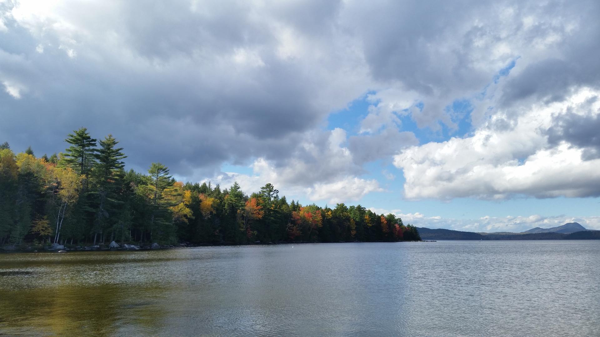 Lake with forested shoreline showing autumn colors under a partly cloudy sky.