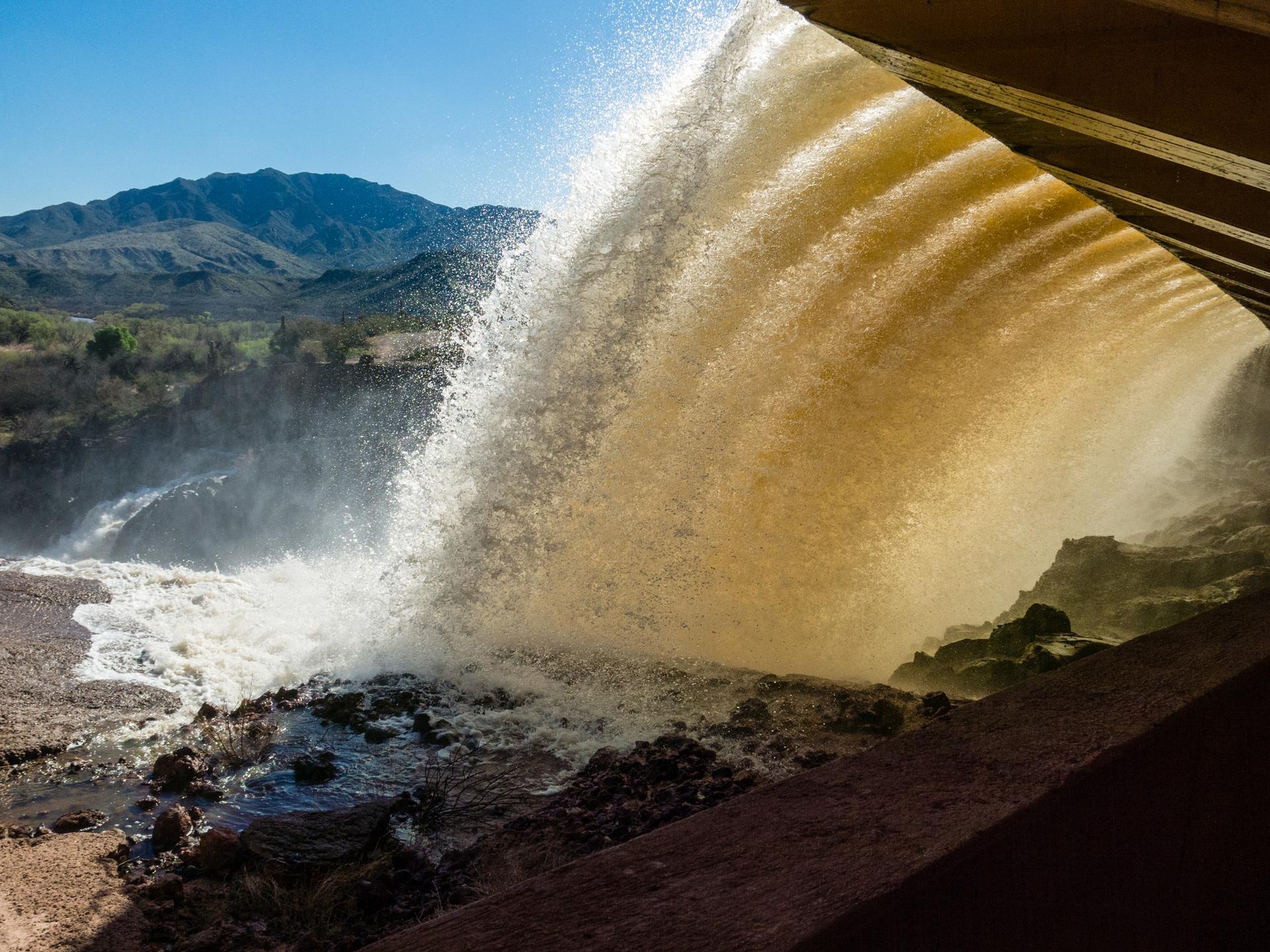 Sunlit brown water powerfully gushes from an outlet, creating mist near distant mountains.