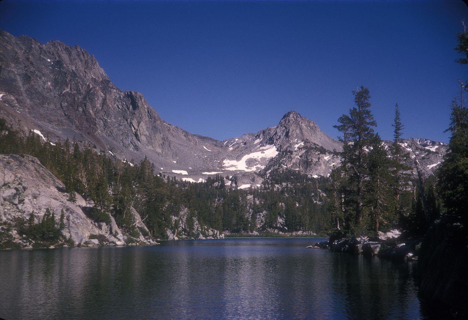 A calm lake reflecting rocky mountains and pine trees under a blue sky.