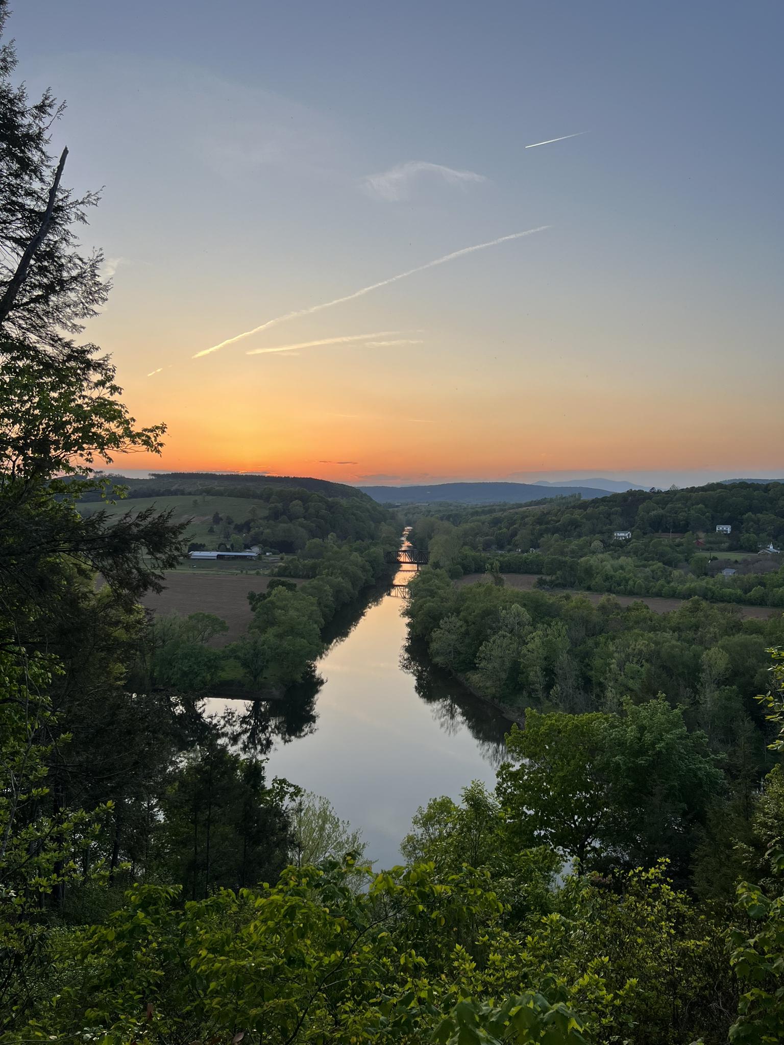 A wide river flows through a valley at sunset, surrounded by green trees.