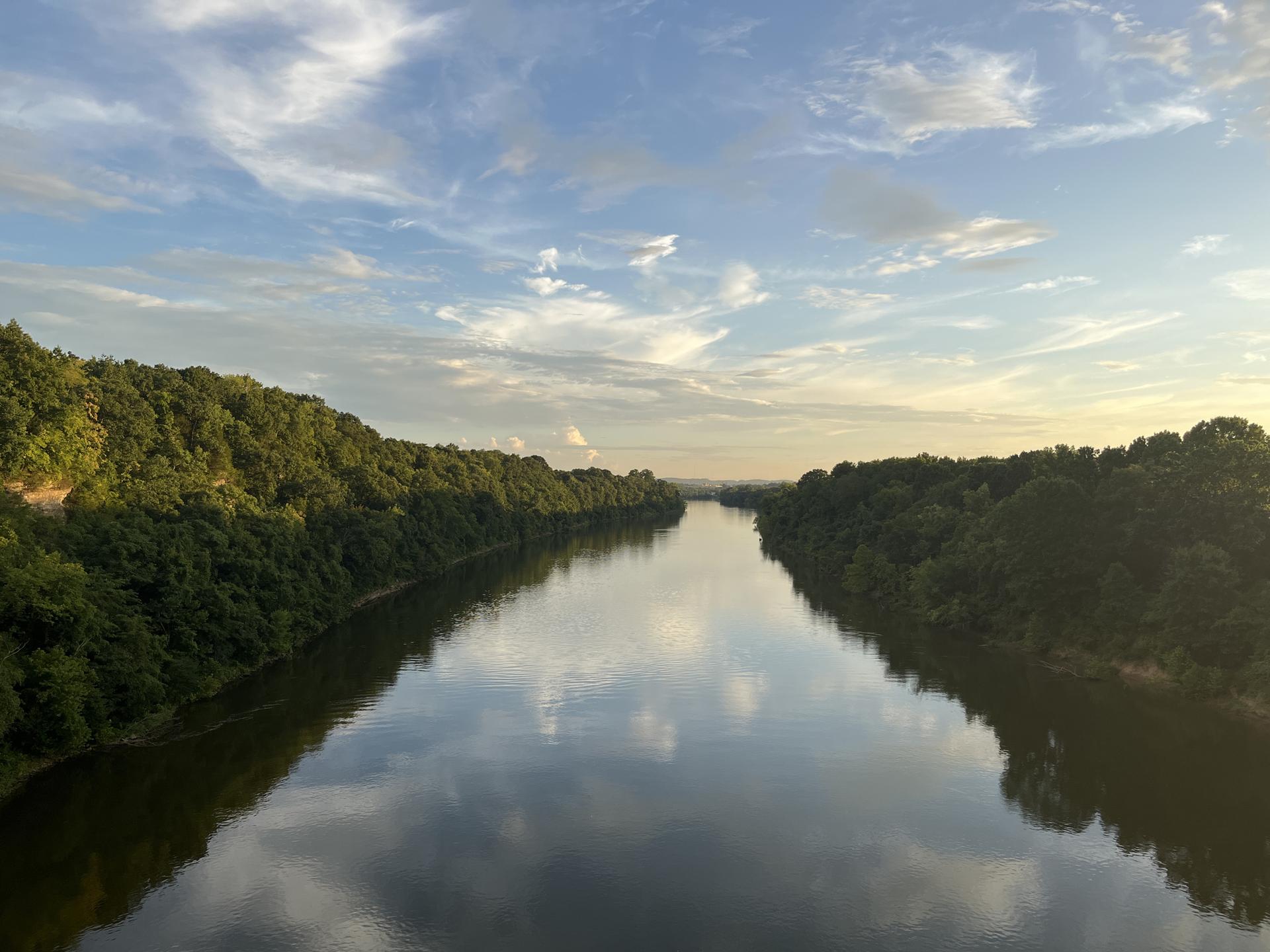 Wide river reflecting cloudy sky, flanked by green forested banks at sunset.